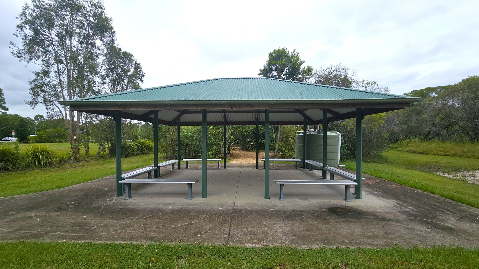 A green-roofed pavilion with open sides and several benches underneath. The structure is supported by green metal poles and stands on a concrete slab. In the background, there are trees and bushes, with a grassy area surrounding the pavilion. A water tank is visible on the right side. The ground is a mix of grass and concrete, and the area appears to be part of a park or recreational space.