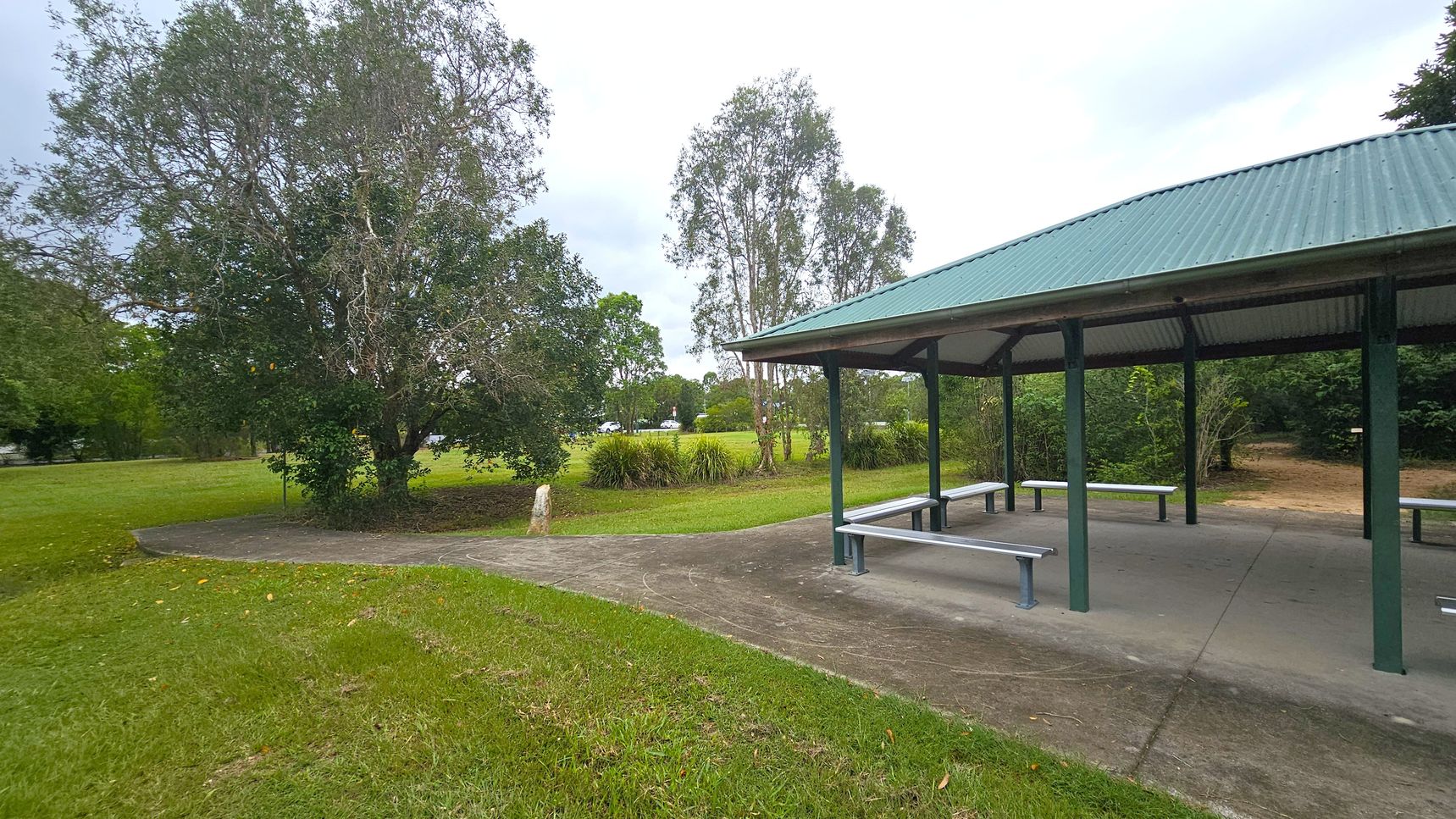 A park scene with a green-roofed shelter on the right, featuring benches underneath. The ground is paved beneath the shelter, transitioning to grass. On the left, there are large trees with dense foliage and a grassy area. In the background, more trees and open grassy fields are visible, with a few shrubs near the shelter. The sky is overcast.