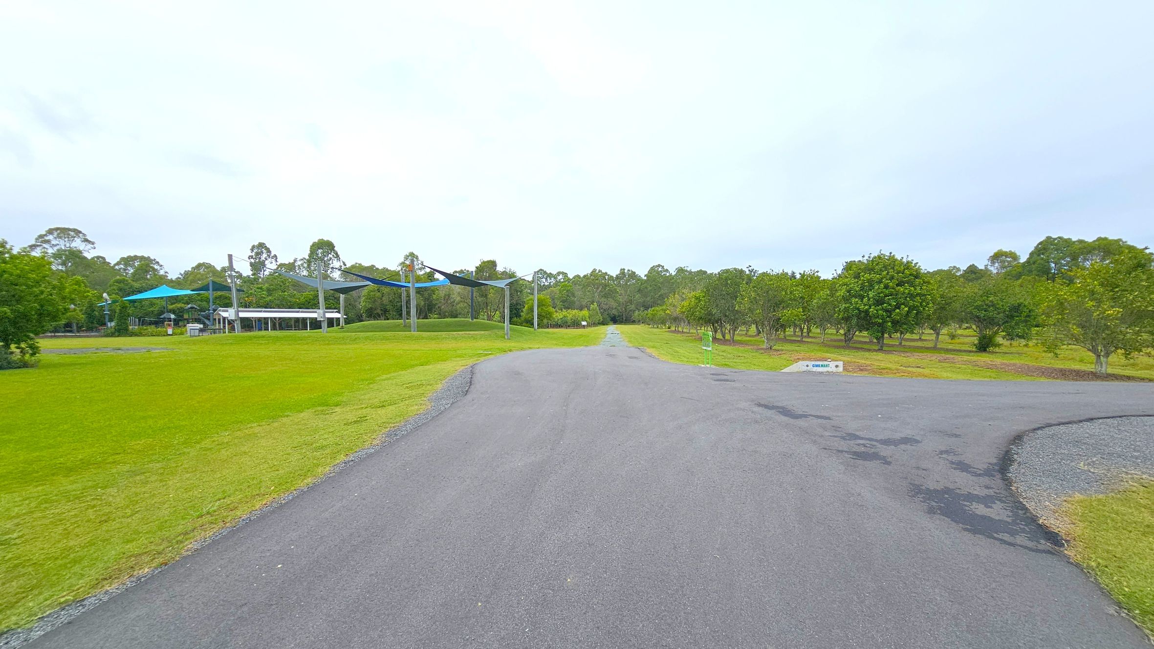 A paved path runs through a park with green grass on either side. On the left, there are blue shade structures and a small building, possibly a picnic area. Trees are scattered throughout the park, with more dense foliage in the background. A sign is visible on the right side of the path. The sky is overcast, and the area appears well-maintained.