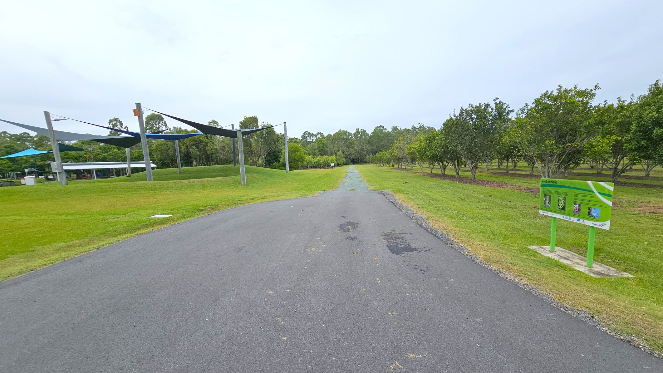 A paved path runs through the centre, flanked by green grass. On the left, there are shade structures with blue and grey canopies supported by poles, and a building in the distance. On the right, a sign with information stands near a row of evenly spaced trees. The background features more trees and a cloudy sky.