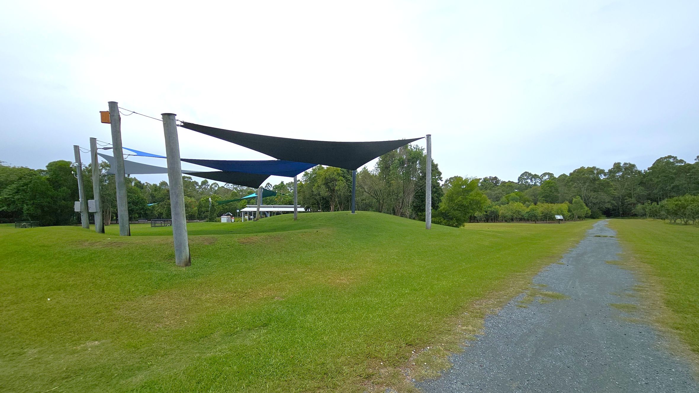A grassy area with several large triangular shade sails supported by tall wooden poles on the left. In the background, there are trees and a small structure with a roof. On the right, a gravel path runs through the grass, leading towards more trees in the distance. The scene is open and spacious, with a mix of green grass and trees.