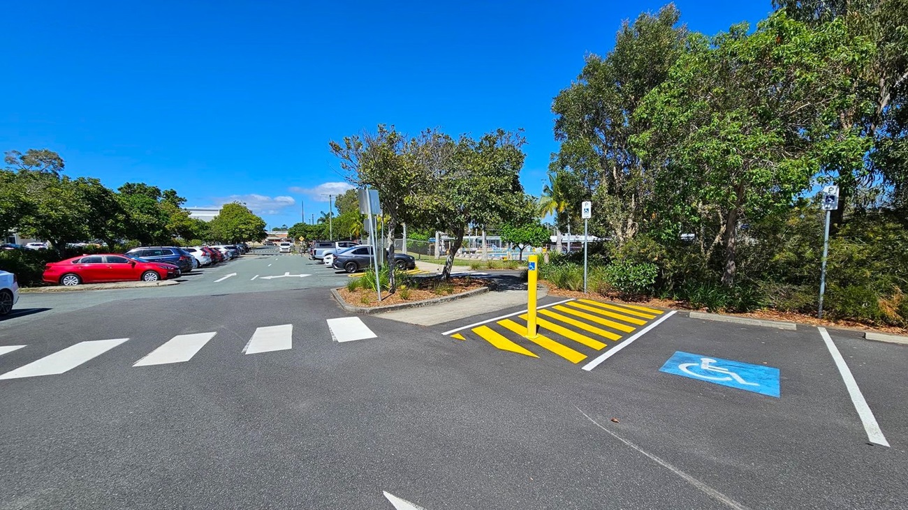 A car park with a pedestrian crossing in the foreground. On the left, there are several parked cars, including a red car. On the right, there is a parking space marked for disabled access with a blue symbol and a yellow-striped area next to it. Trees and shrubs line both sides of the car park, providing greenery. In the background, more parked cars and additional trees are visible.