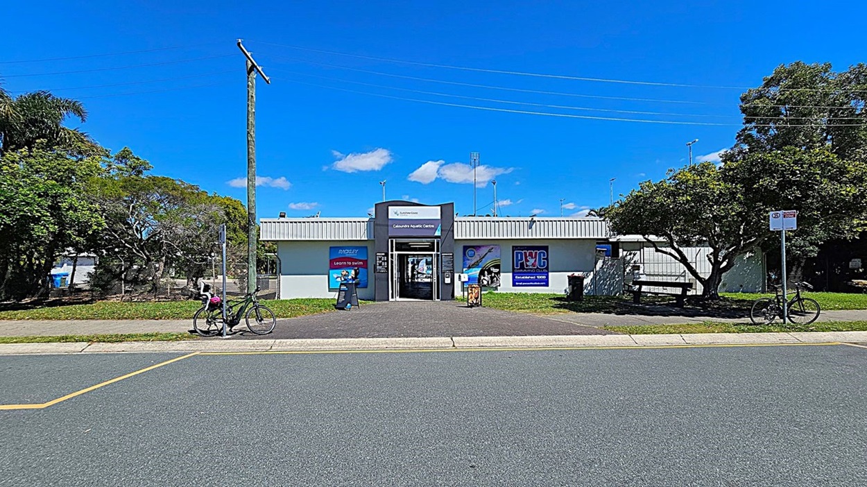 A single-storey building with a grey and white facade and a sign reading "Caloundra Aquatic Centre" above the entrance. The entrance is flanked by promotional posters. In front of the building, there is a paved pathway leading to the entrance, with a bicycle parked on the left side and another on the right side near a bench and a tree. The building is surrounded by greenery, including trees and bushes, with a power pole on the left side. The road in the foreground has a marked parking area.