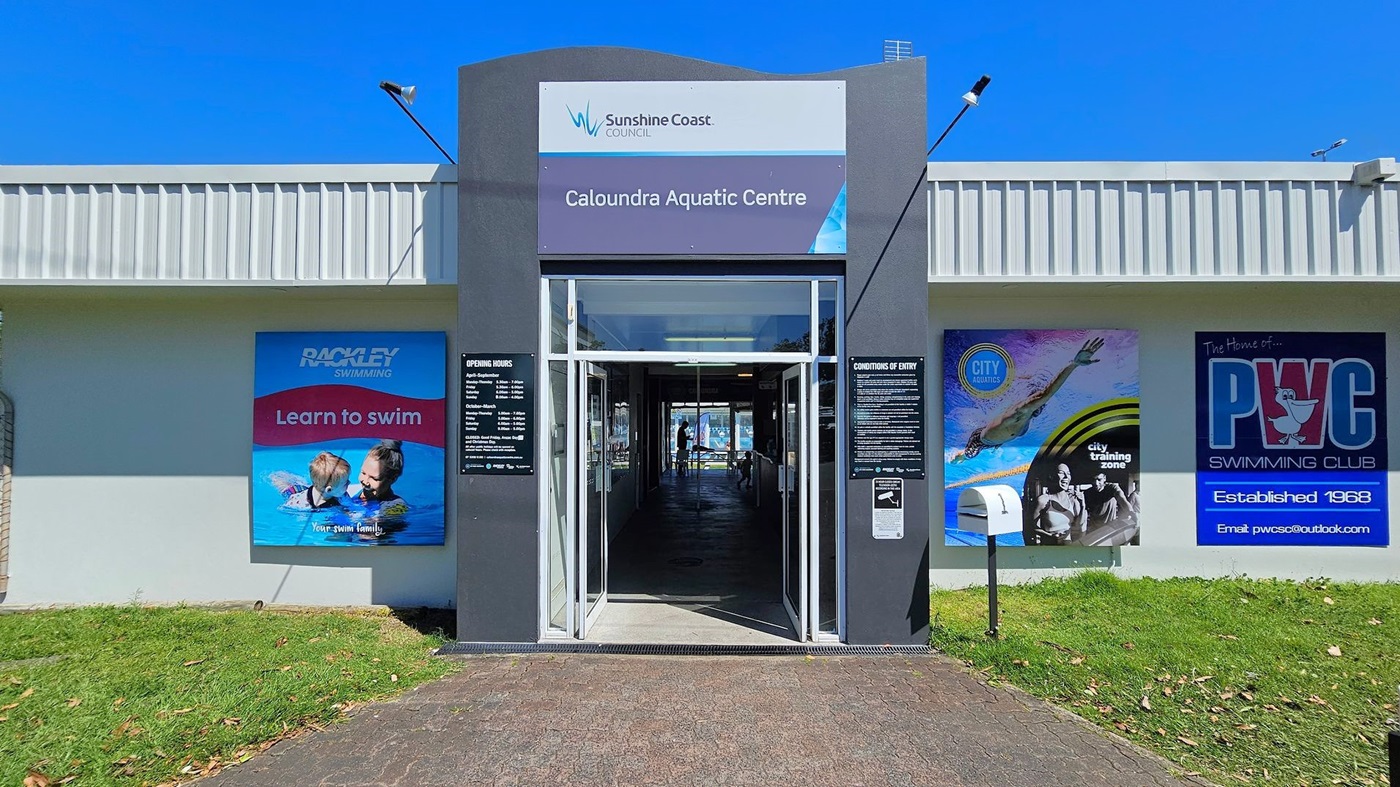 Entrance to the Caloundra Aquatic Centre with a sign above the door displaying the name and Sunshine Coast Council logo. On the left, a poster for Rackley Swimming promoting "Learn to swim" with an image of an adult and child in a pool. On the right, a poster for PWC Swimming Club, established in 1968, and another poster for City Aquatics. The ground is paved with a grassy area on either side. The building has a corrugated metal roof.