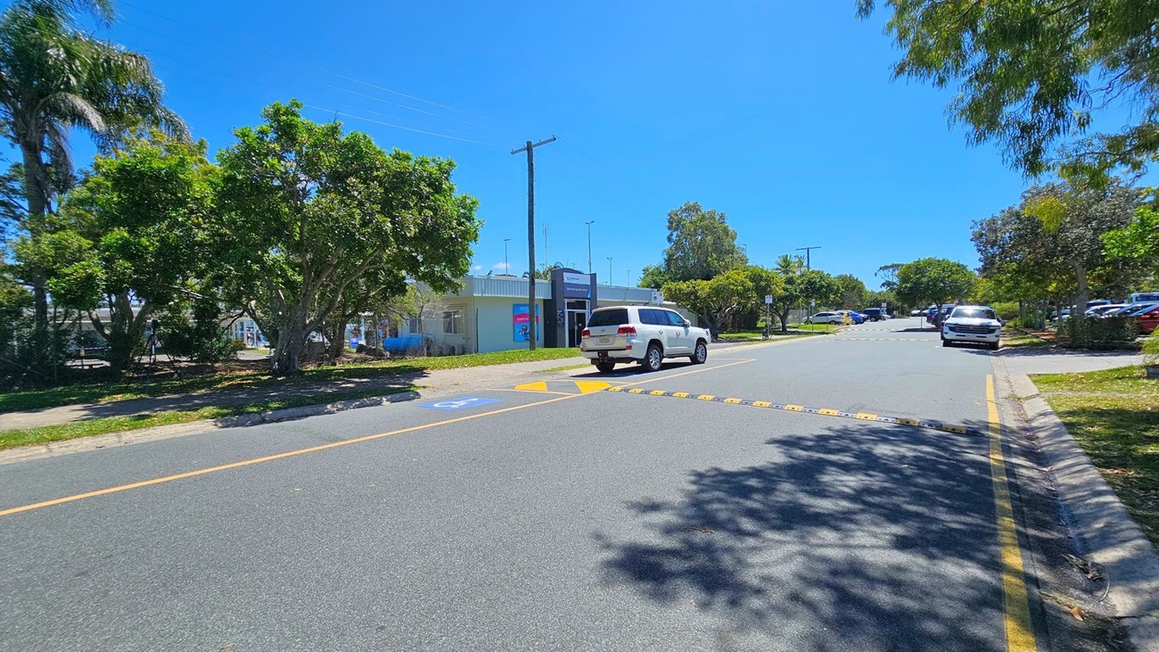 A street with a speed bump in the centre and a white SUV parked on the right side. On the left, there are large green trees and a building with a sign and a blue poster. The pavement is visible on both sides of the road, with shadows from the trees cast on the ground. Further down the street, more parked cars and additional trees are visible. The sky is clear and blue.