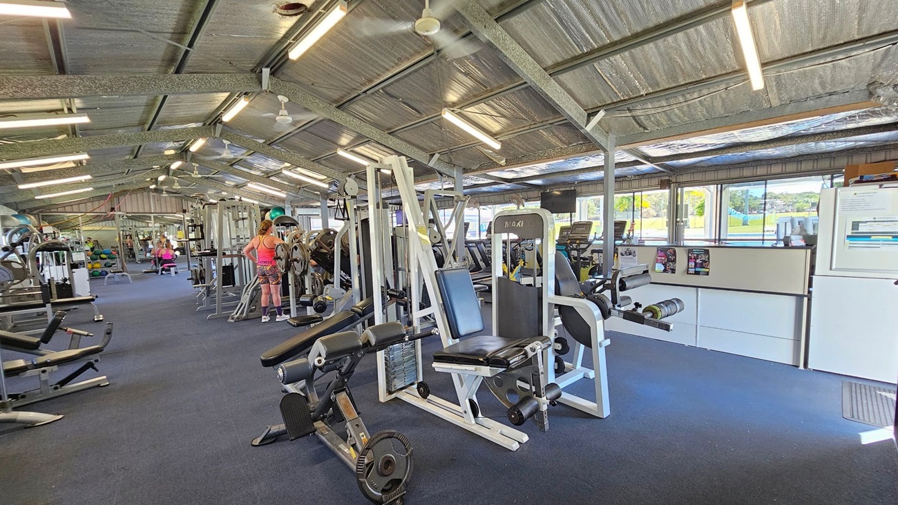 A gym interior with various exercise machines and equipment. On the left, there are weight machines and benches. In the middle, a person in colourful workout attire stands near a machine. On the right, there are more machines, a white counter with posters, and large windows showing an outdoor view. The floor is carpeted in dark blue, and the ceiling is metallic with exposed beams and fluorescent lights.