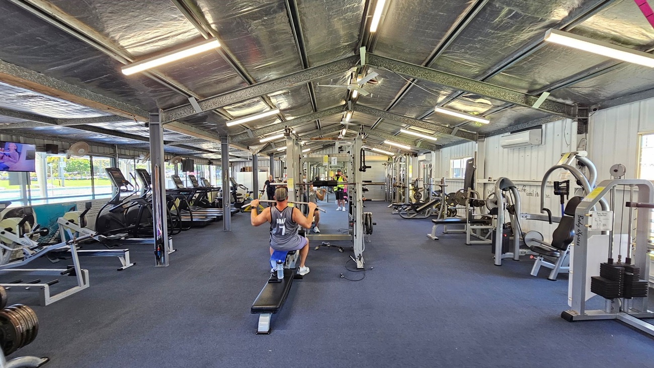 A gym with various exercise equipment. On the left, there are several treadmills and rowing machines lined up against the wall, with a television mounted above them. In the middle, a person is using a lat pulldown machine, facing away from the camera. On the right, there are weight machines and benches. The floor is carpeted in blue, and the ceiling is metallic with exposed beams and fluorescent lights. The walls have windows and air conditioning units.