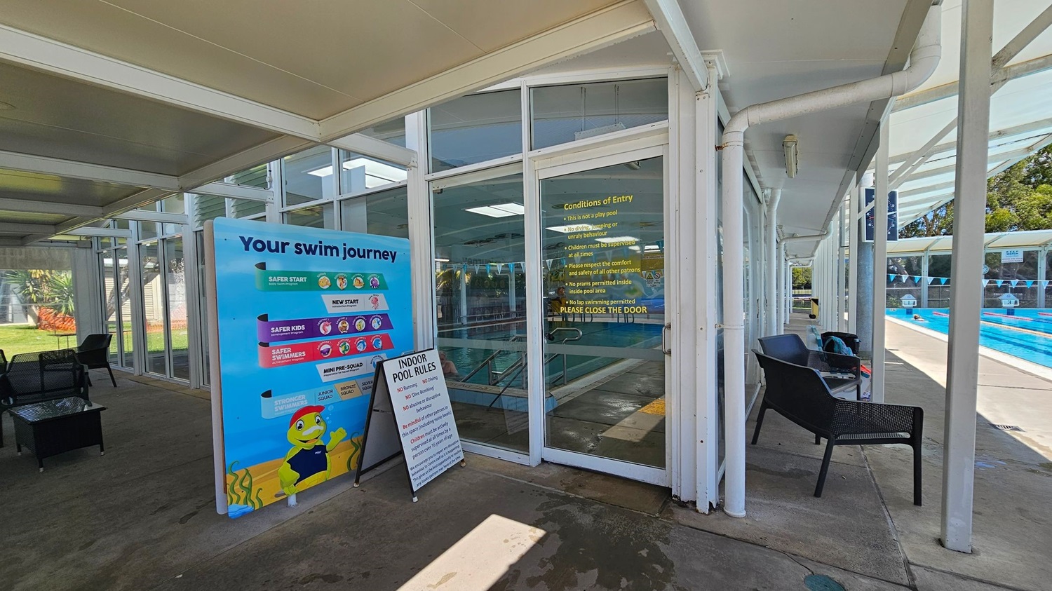 A swimming pool area with a glass entrance door on the right, displaying "Conditions of Entry" in yellow text. To the left of the door, a colourful sign titled "Your swim journey" outlines different swimming levels. In front of the sign, a smaller board lists "Indoor Pool Rules." Black chairs are placed along the glass wall, and the pool with lane markers is visible through the glass. The floor is concrete, and the ceiling is white with exposed beams.