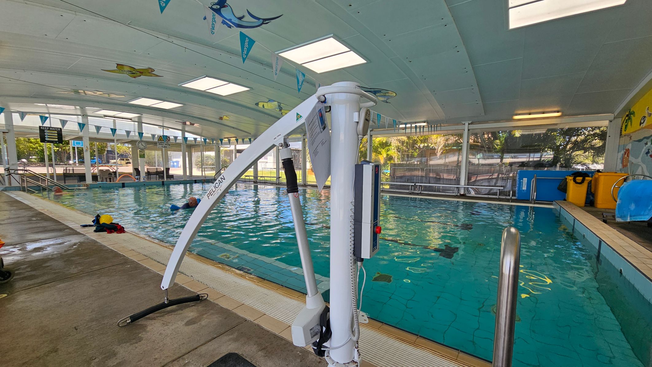 Indoor swimming pool with a hoist on the right side. The pool has clear water and a tiled edge. On the left, there is a wet concrete floor with some towels and swimming gear. The ceiling is white with skylights and decorated with aquatic-themed banners. The background shows large windows with views of greenery and outdoor seating. There are also colourful bins and a mural on the far right wall.