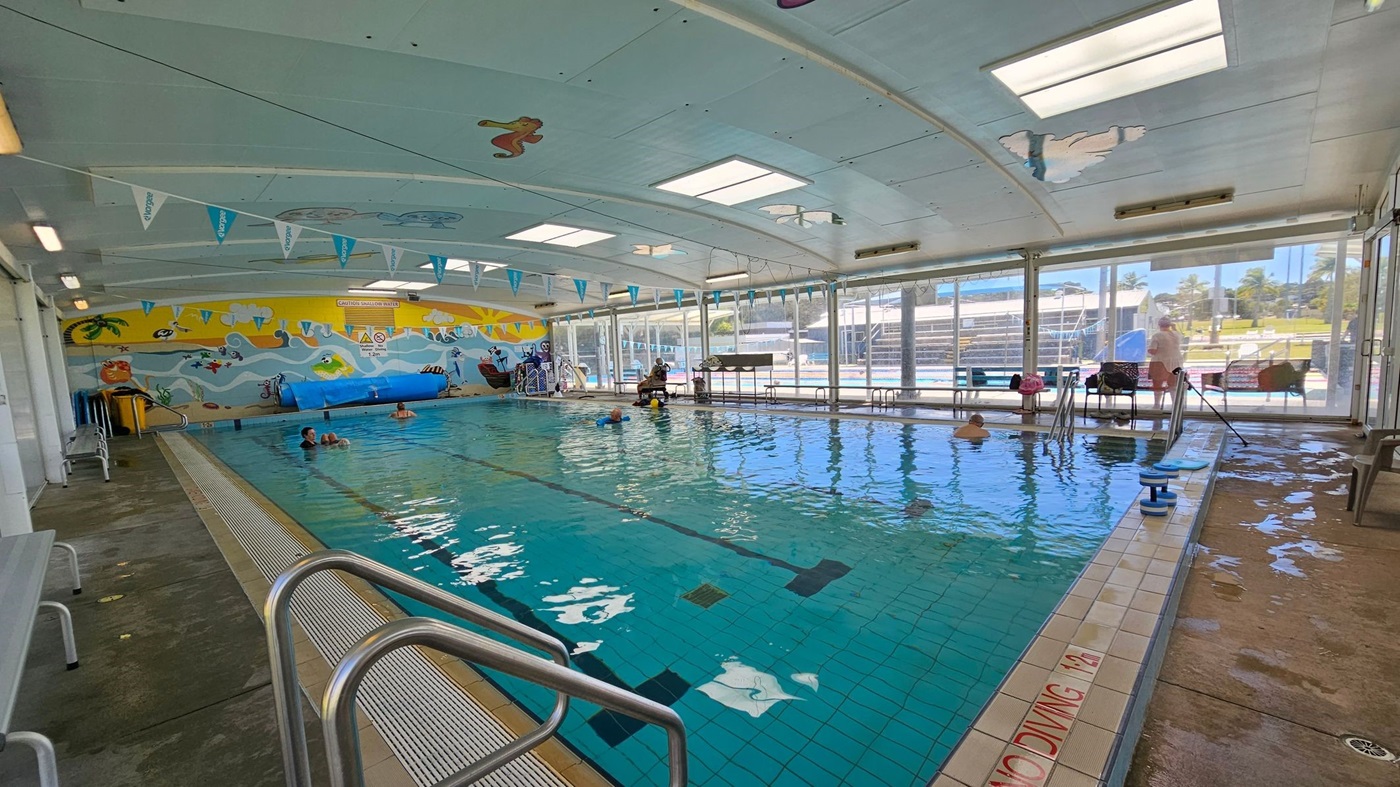 Indoor swimming pool with a tiled floor and a ceiling featuring skylights. The left side has a bench and a colourful mural with sea creatures and a pirate theme. The right side has large windows showing an outdoor area with greenery and a playground. Several people are in the pool, and there are poolside chairs and equipment. The foreground has metal handrails leading into the pool. Triangular flags hang from the ceiling.