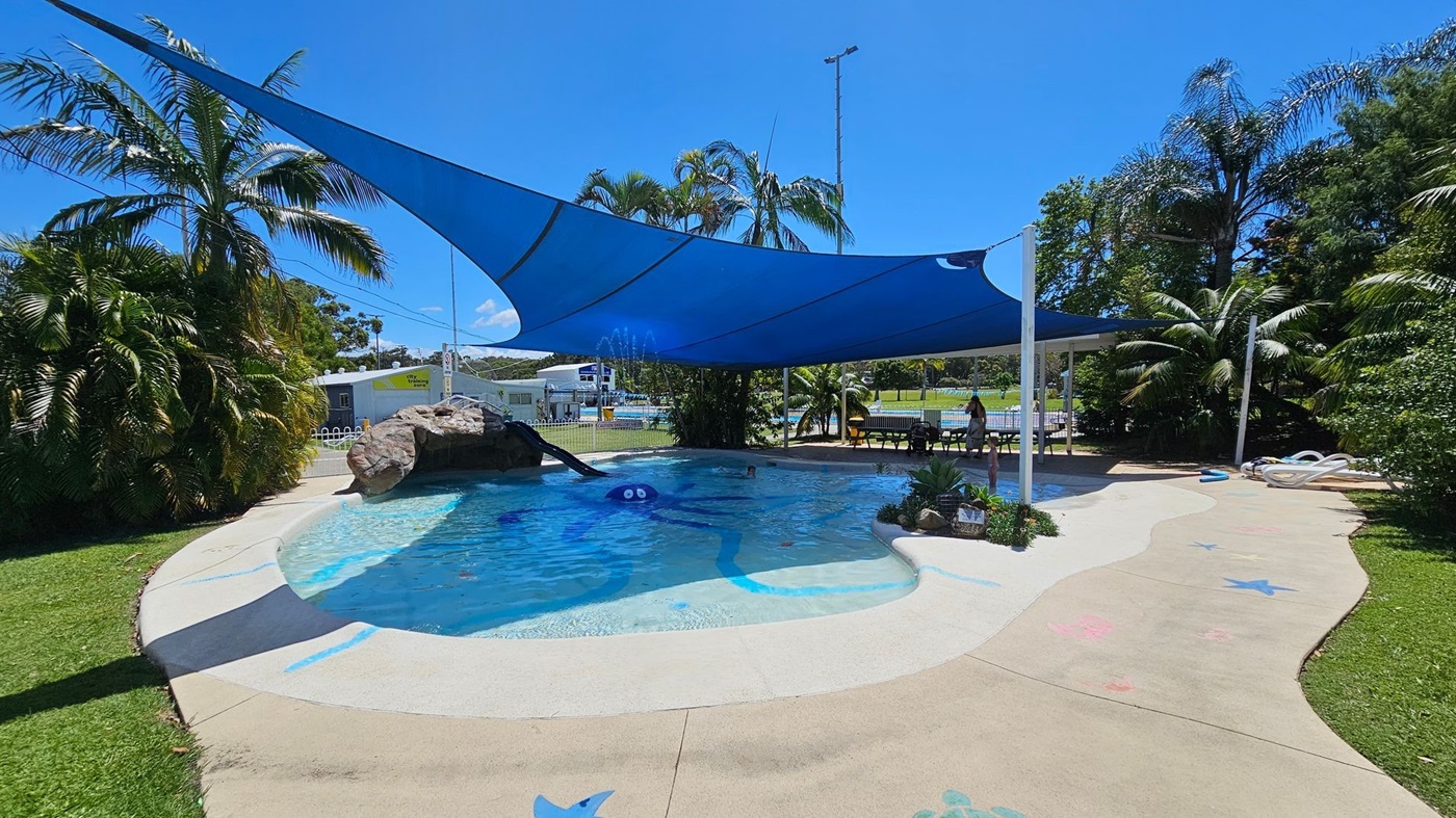 A poolside café with a counter on the right displaying a blender, jars of snacks, and a sign reading "POOL DECK CAFÉ." On the left, there are ice cream freezers with "Peters" branding and a display of ice cream options. In the middle, a gate with signs, including one that says "KEEP WATCH" and another with a cartoon duck and pool rules. Beyond the gate, there are blue shade sails, palm trees, and a white picket fence. The floor is carpeted, and the ceiling is white.
