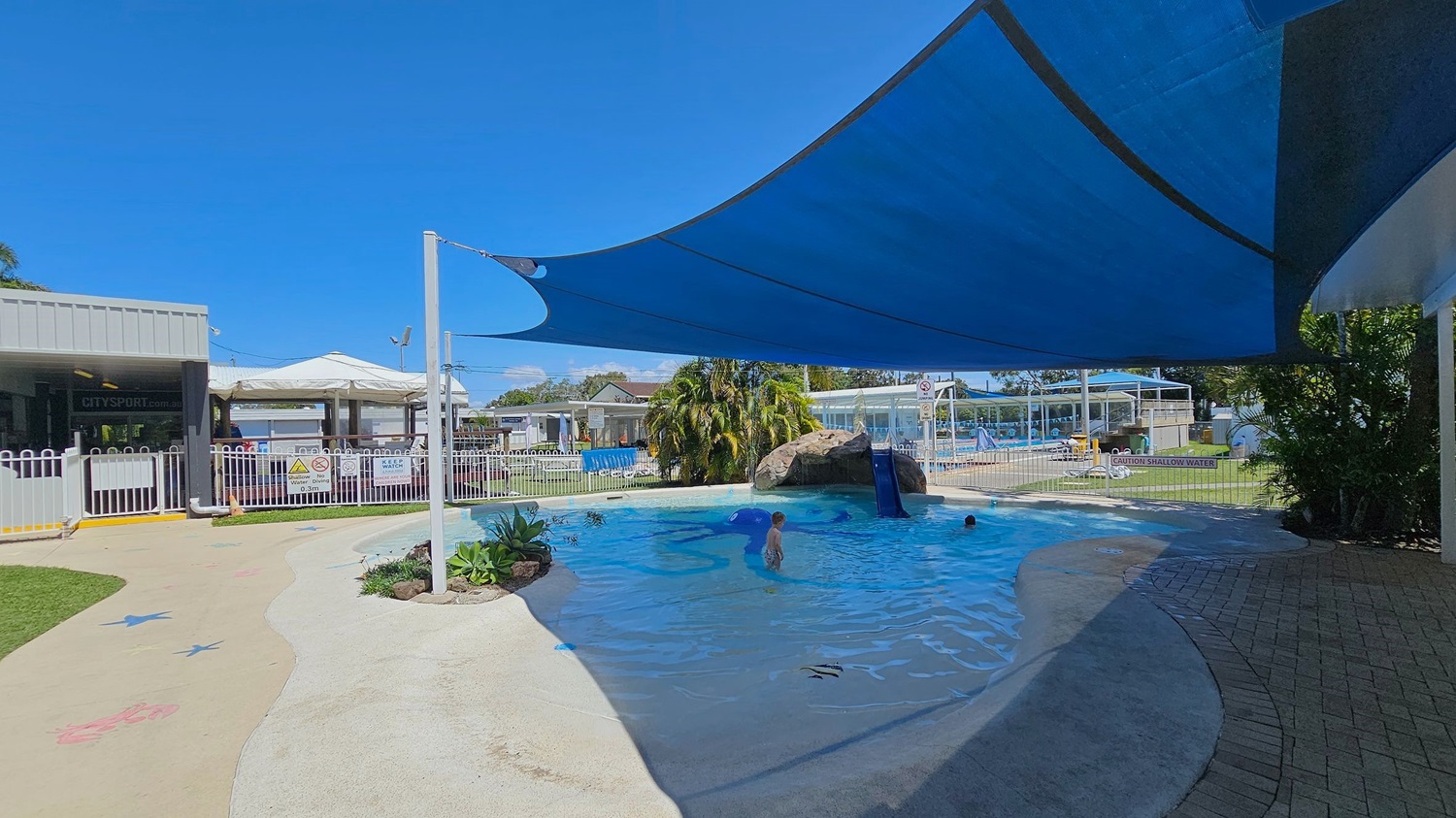 A shallow outdoor pool with a blue shade sail overhead. On the left, there is a fenced area with a sign that reads "City Sport" and a white gate. The pool has a small slide attached to a rock structure in the middle. There are a few children playing in the water. The ground around the pool is a mix of concrete and brick paving. In the background, there are more fenced areas, a grassy lawn, and some buildings with white roofs.