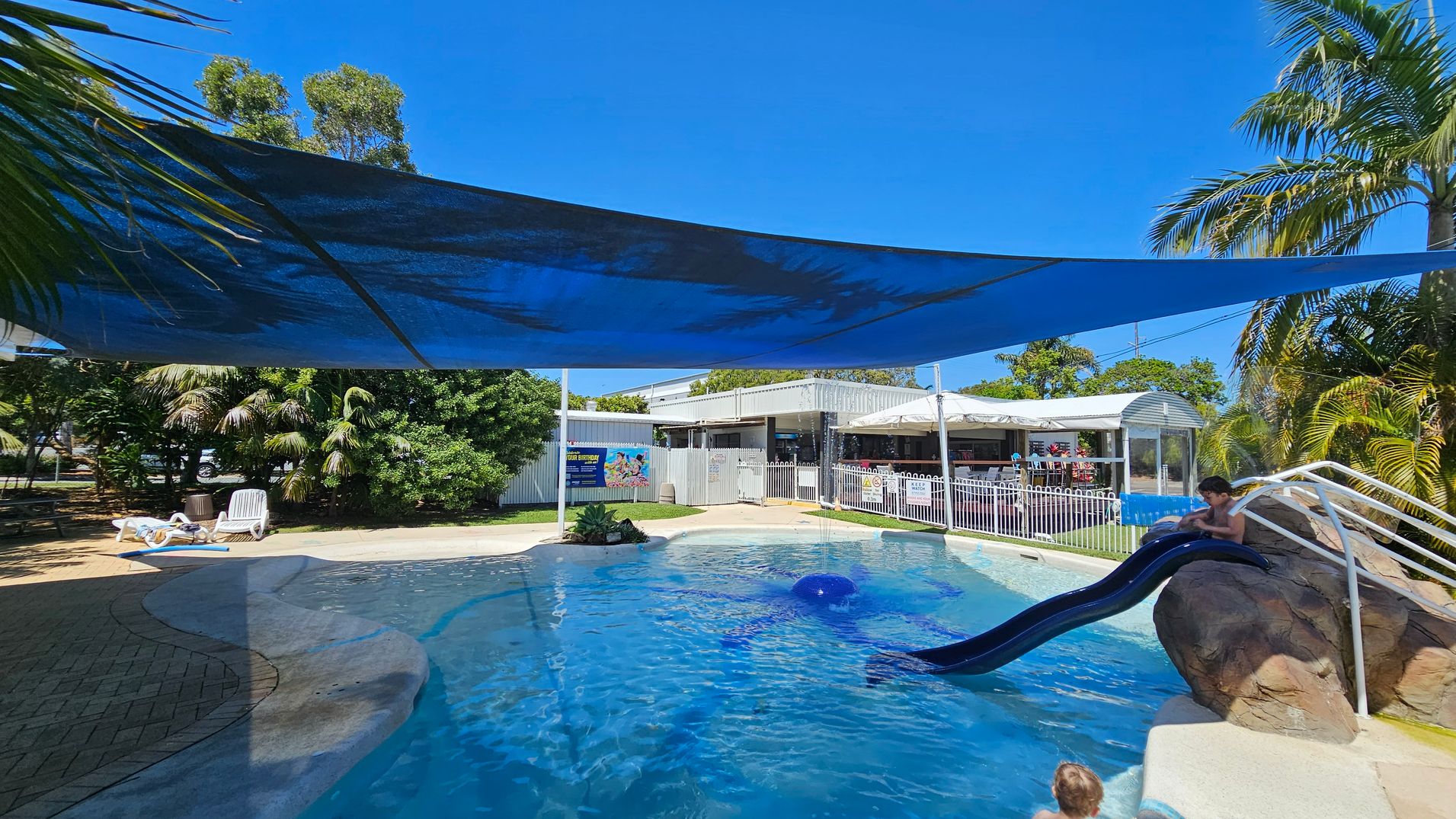 A swimming pool area with a blue shade cloth overhead. On the right, a child is sitting at the top of a blue slide attached to a rock formation. In the middle, the pool water is clear with a small fountain feature. On the left, there are white lounge chairs on a paved area, surrounded by lush green trees and plants. In the background, there is a white fence and a building with a covered patio area.