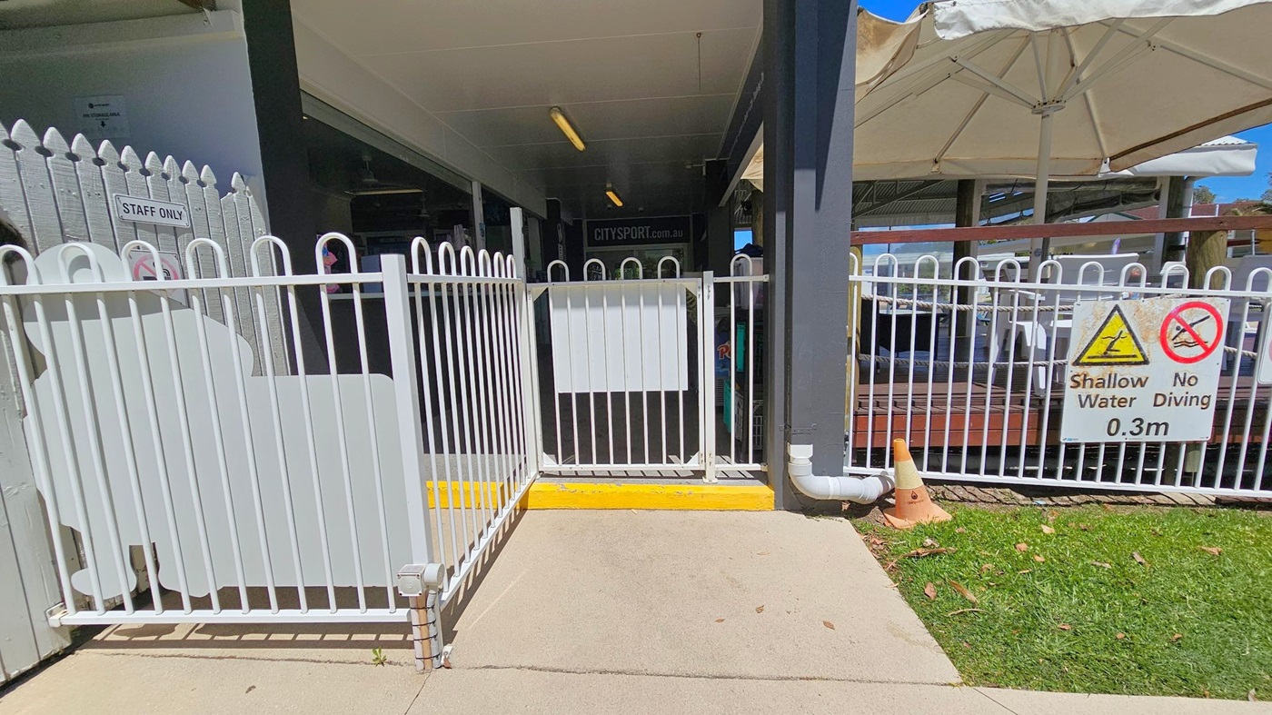 White metal fence with a gate in the centre. On the left, a "Staff Only" sign is attached to the fence. On the right, a sign warns of shallow water (0.3m) and no diving. Behind the fence, a shaded area with a white umbrella and seating is visible. The ground is a mix of concrete and grass, with a small orange traffic cone near the right side. The ceiling is covered by a canopy.