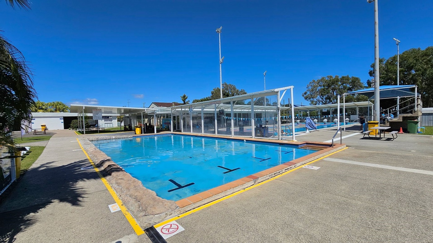 An outdoor swimming pool area with two pools. The pool on the right is smaller and has a rocky edge, while the pool on the left is larger and covered with a transparent roof. The ground around the pools is paved with a mix of green and beige surfaces, marked with yellow lines and warning signs. In the background, there are tall light poles, grassy areas, palm trees, and a few benches. A person is standing near the pool on the right, and some white lounge chairs are visible.