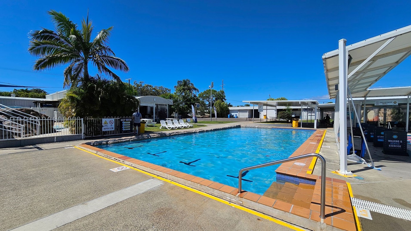 Outdoor swimming pool with clear blue water. On the left, there is a white fence, a palm tree, and several white sun loungers on a grassy area. In the middle, the pool is bordered by a brick edge and a metal handrail leading into the water. On the right, there is a covered area with a white roof and signs indicating "Walking & Gentle Exercise." The ground around the pool is a mix of concrete and textured surfaces.