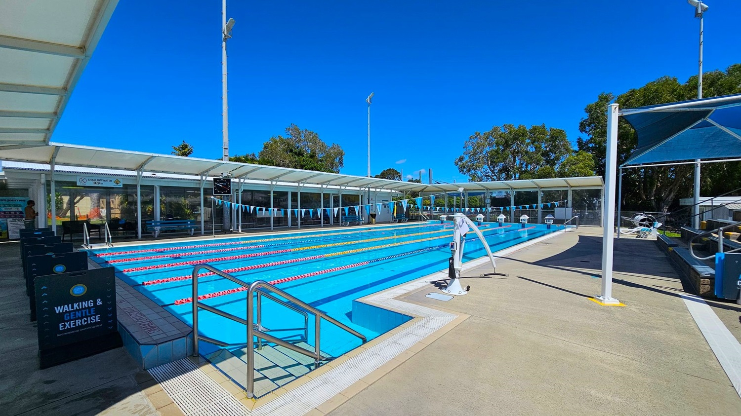 An outdoor swimming pool with multiple lanes, each separated by floating lane markers. On the left, there is a covered walkway with signs indicating "Walking & Gentle Exercise" and "Shallow Water 0.9m." The middle of the image shows the pool with blue water and lane dividers in red, yellow, and white. On the right, there is a pool lift and shaded seating area. In the background, there are trees and a clear blue sky. The floor around the pool is a mix of concrete and non-slip surfaces.