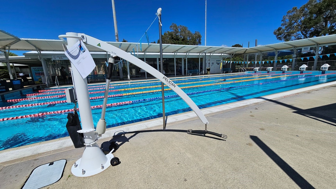A swimming pool with multiple lanes separated by floating lane markers in red, yellow, and blue. On the left, there is a white pool lift labelled "PELICAN" with a control panel and a hanging document. The pool area is surrounded by a covered walkway with transparent roofing. In the background, there are trees and a building with large windows. The floor around the pool is a light grey concrete surface.