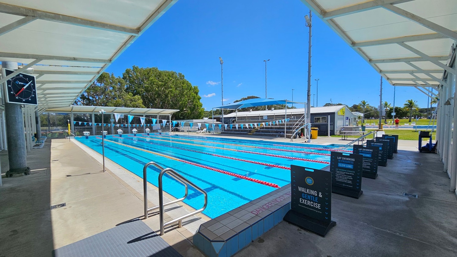 An outdoor swimming pool with multiple lanes, each separated by red and yellow lane dividers. On the left, a large clock is mounted on a column under a white canopy. The pool deck is concrete, with a metal handrail leading into the water. On the right, several signs provide information about lap swimming and exercise. In the background, there are trees, a small building, and a grassy area with palm trees. The sky is clear and blue.