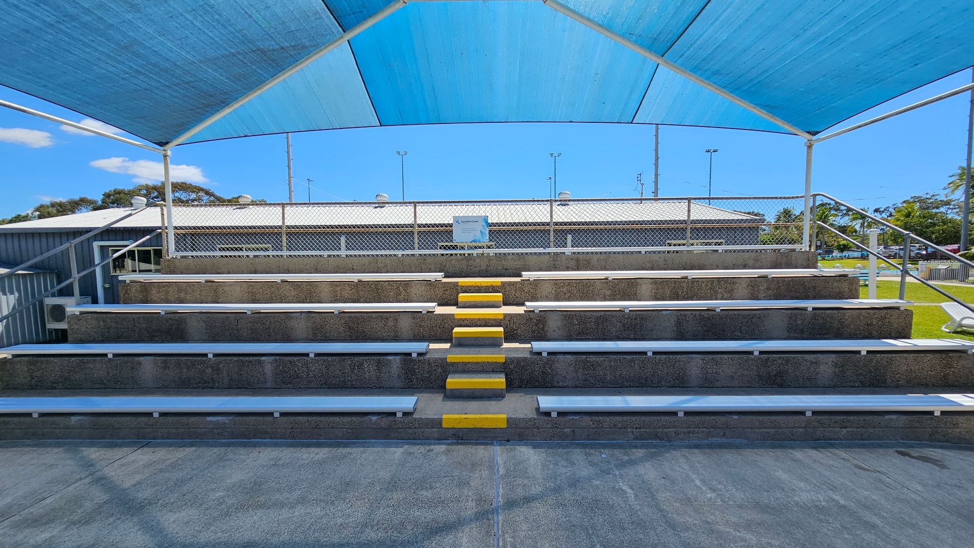 Concrete bleachers with metal seating and yellow-painted steps in the centre. A blue shade cloth is stretched overhead, supported by white metal poles. A chain-link fence is behind the bleachers, with a building and some trees visible in the background. The ground in front of the bleachers is paved with concrete.