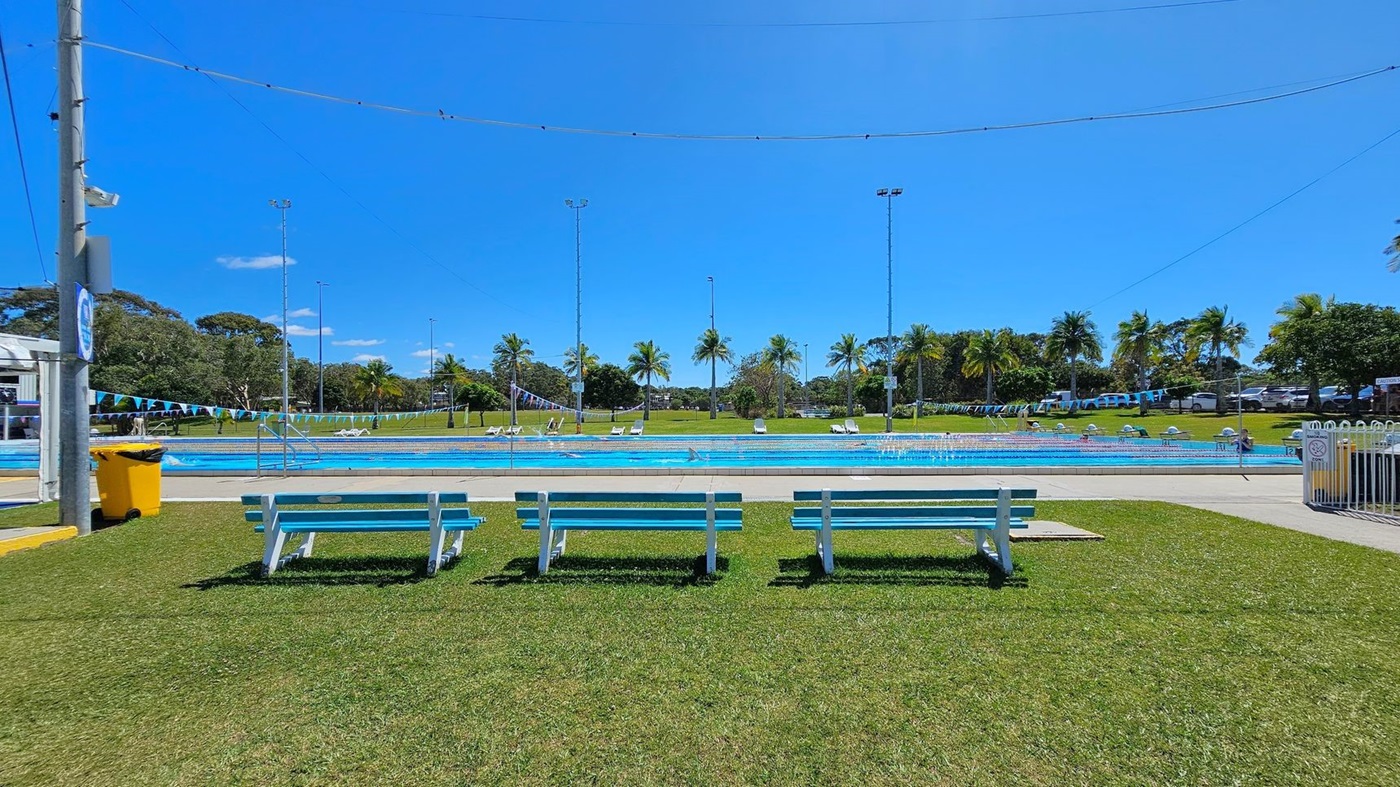 Three white and blue benches are positioned on a grassy area in the foreground. Behind the benches, there is a swimming pool with lane dividers and a few people lounging on poolside chairs. Palm trees and other greenery are visible in the background, along with several tall light poles. A yellow rubbish bin is on the left side near a pole, and a white fence is on the right side. The ground around the pool is paved.