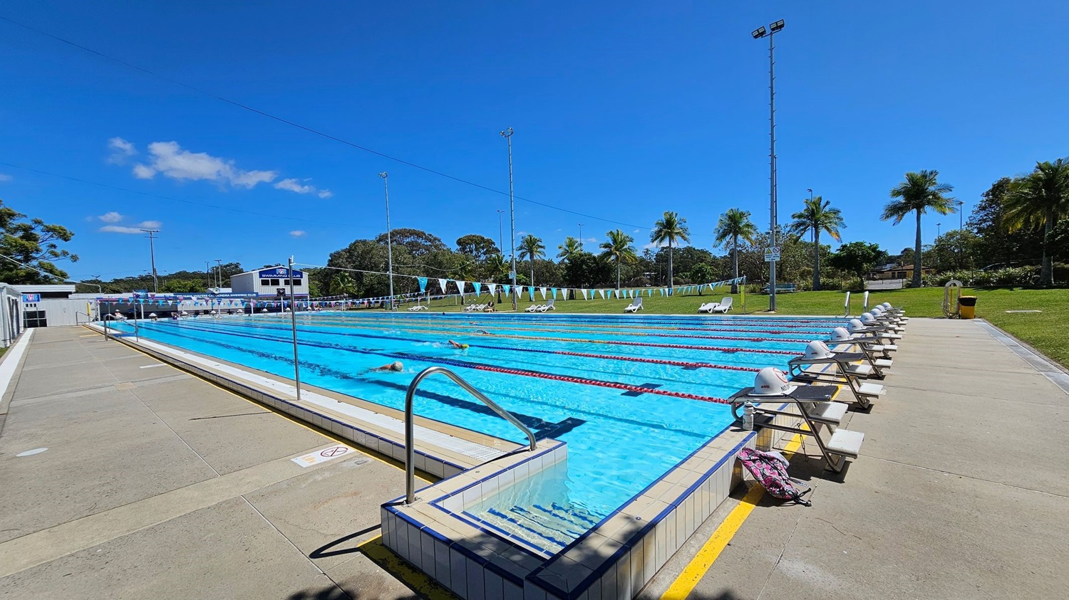 An outdoor swimming pool with multiple lanes, each marked by lane ropes. On the left, there is a concrete walkway with a metal handrail leading into the pool. On the right, there are starting blocks lined up along the pool's edge, with a grassy area and palm trees in the background. A few white lounge chairs are scattered on the grass. In the middle, swimmers are practising in the lanes. The background features a scoreboard and some buildings.