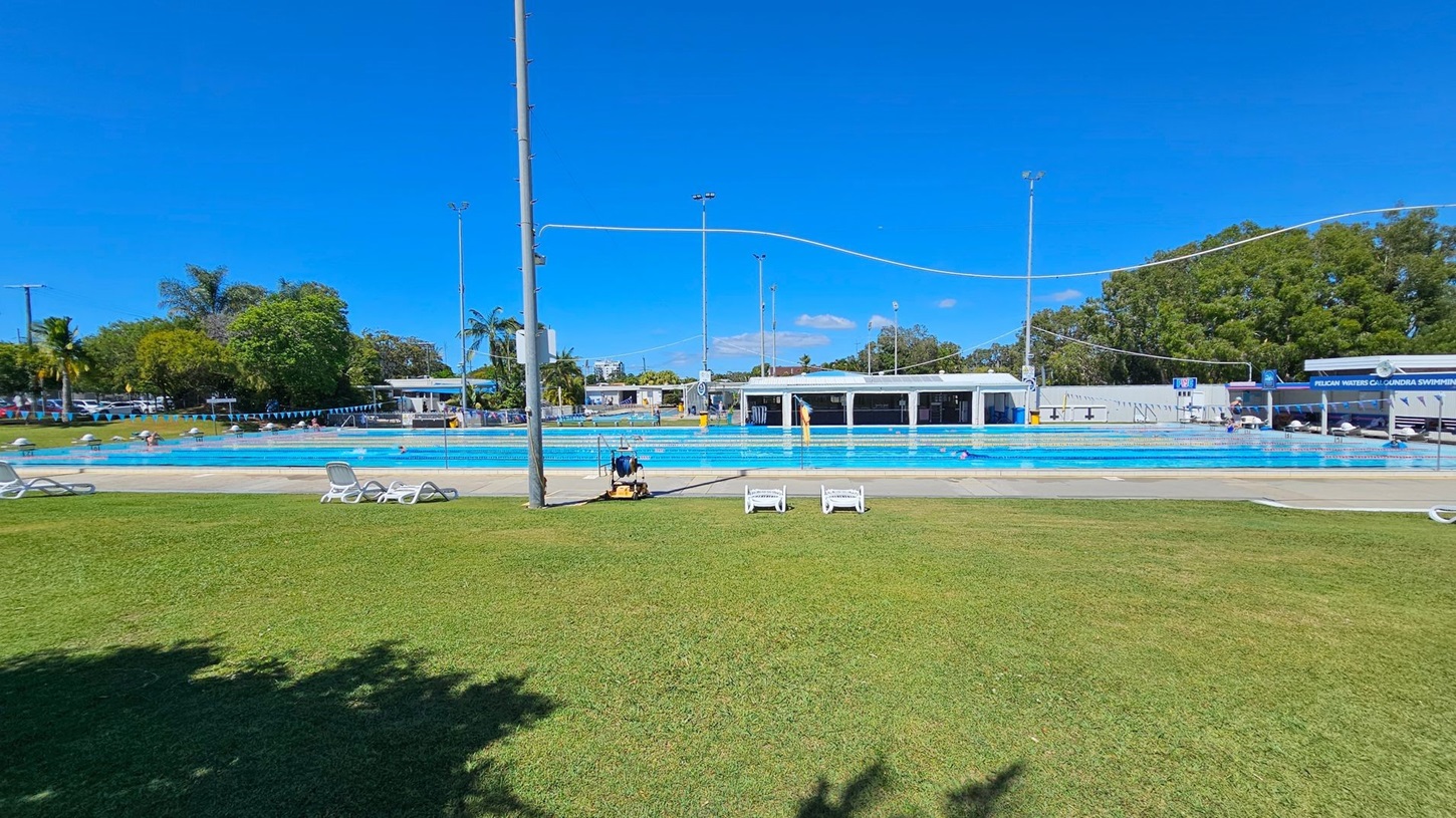 A large outdoor swimming pool with blue water is in the centre, surrounded by a concrete deck. On the left, there are trees and parked cars. In the middle, a person is sitting on a chair near the pool, and there are two white lounge chairs on the grass. On the right, there is a building with a sign that reads "Pelican Waters Caloundra Swimming." The background features more trees and some buildings. The foreground has a well-maintained grassy area.