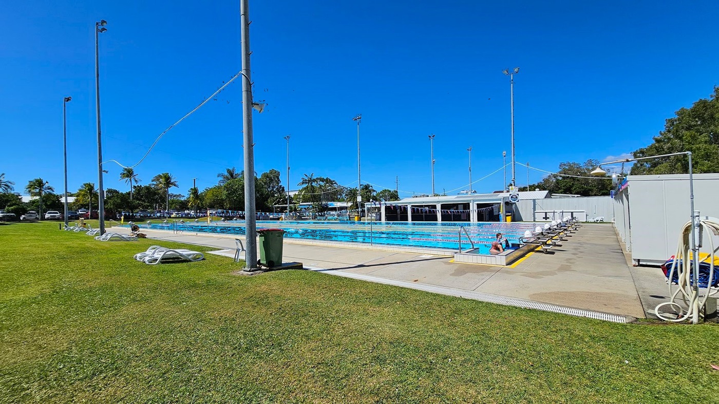 Outdoor swimming pool with lanes in the centre, surrounded by a concrete deck. On the left, there is a grassy area with several white lounge chairs and tall light poles. In the background, there are palm trees and parked cars. On the right, there are more lounge chairs, a shower area, and a building with a shaded area. The pool has lane dividers and a few people swimming. The sky is clear and blue.