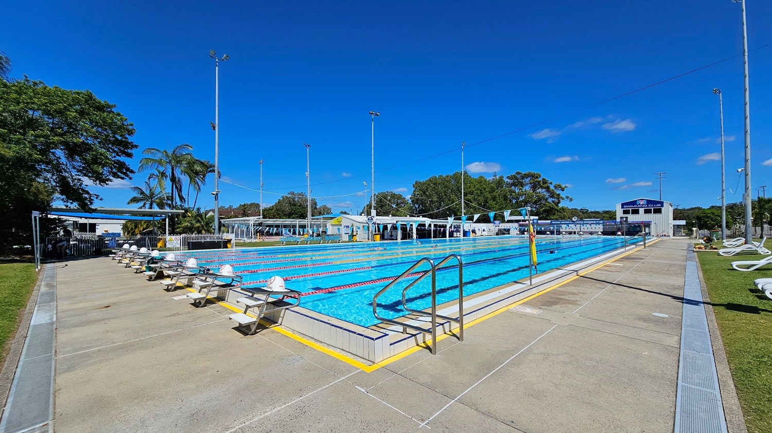 An outdoor swimming pool with multiple lanes, each marked by lane ropes. Starting blocks are positioned at the near end of the pool. On the left, there is a grassy area with trees and a shaded seating area. On the right, there are white lounge chairs on a grassy strip. In the background, there are buildings, including a structure with a sign that reads "Swim Club," and tall light poles surrounding the pool area. The pool deck is concrete.