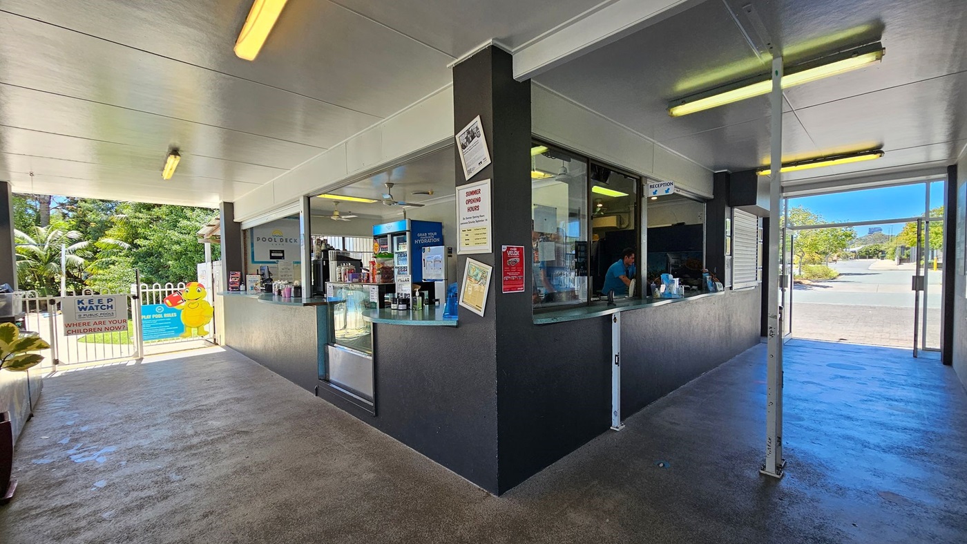 Entrance to the Caloundra Aquatic Centre. The entrance is flanked by two dark grey pillars with a sign above reading "Sunshine Coast Council Caloundra Aquatic Centre." On the left, a poster for Rackley Swimming with the text "Learn to swim" and opening hours listed below. On the right, a poster for City Aquatics and PWC Swimming Club. The ground is paved, and there is grass with scattered leaves in front of the building. The ceiling inside the entrance is white with fluorescent lighting.