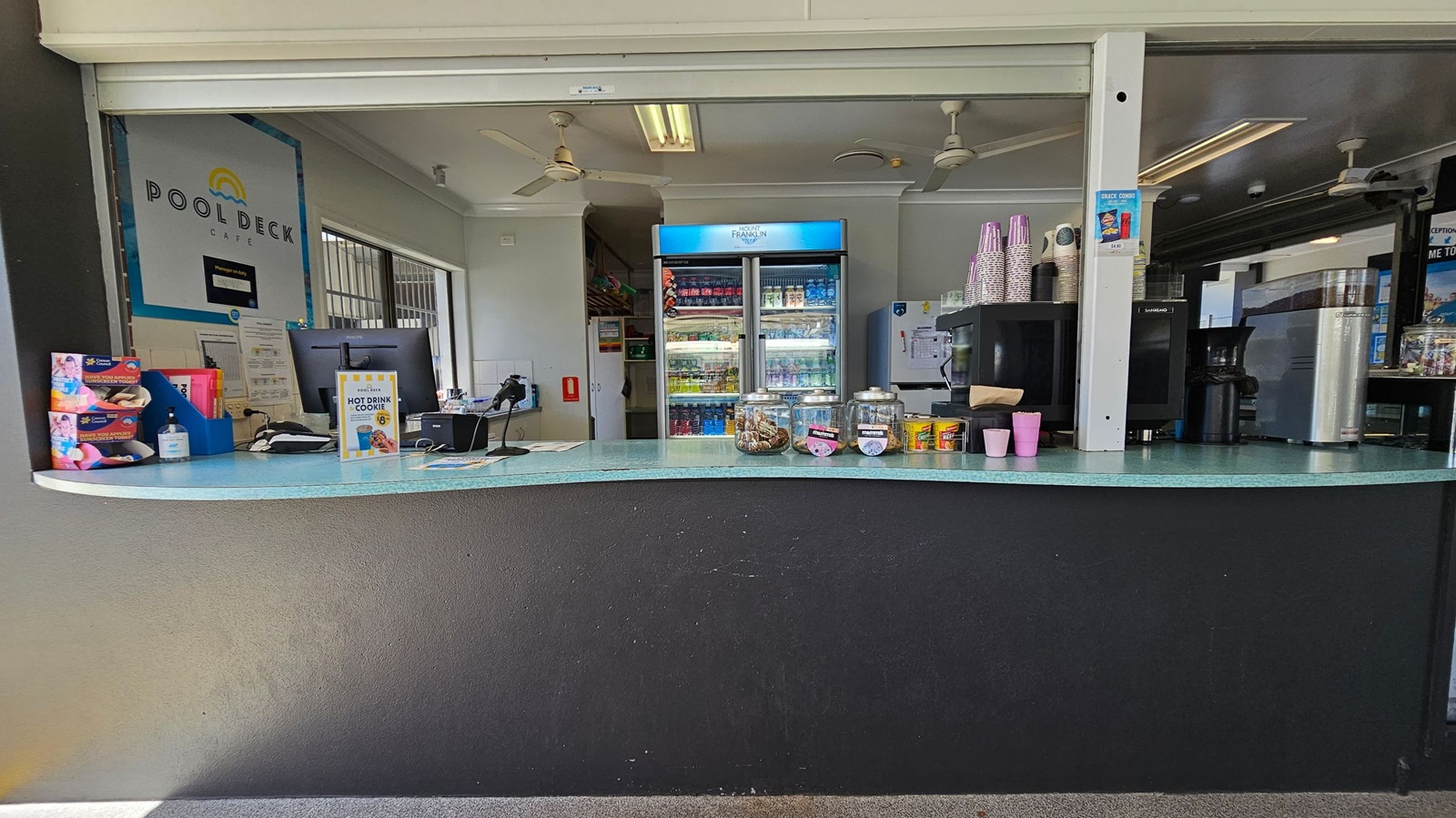 A café counter with a turquoise surface and a black front. On the left, there are boxes of snacks, a hand sanitiser bottle, and a computer monitor. In the middle, there are jars with cookies and a sign advertising a hot drink and cookie deal. On the right, there are stacks of paper cups, a coffee machine, and a refrigerator filled with drinks. The background has a "Pool Deck Café" sign, a ceiling with fans, and fluorescent lights. The floor is carpeted.