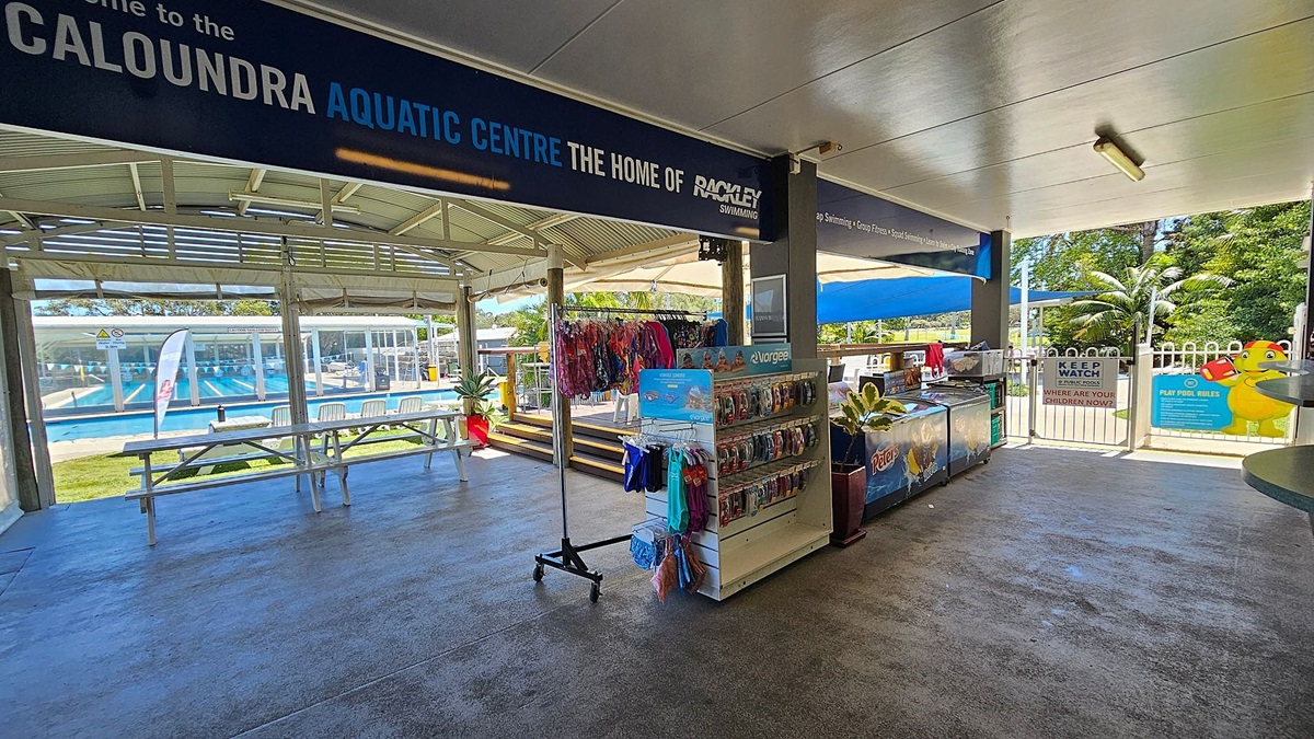 A covered area with a sign reading "Caloundra Aquatic Centre" at the top. On the left, there are picnic tables and a view of a swimming pool with lanes. In the middle, there is a rack displaying swimwear and accessories. On the right, there is a counter with a freezer and a sign that says "Keep Watch Around Pools." The floor is concrete, and the ceiling is white with fluorescent lighting. In the background, there are trees and a fence with a gate.