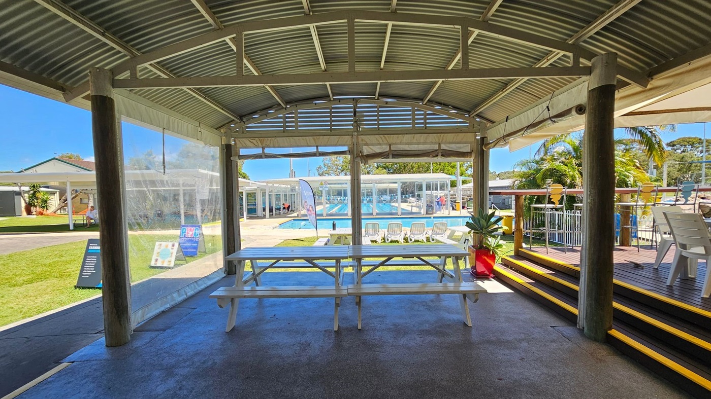 A covered outdoor area with a corrugated metal roof and wooden support beams. In the foreground, there are white picnic tables on a concrete floor. To the right, there are steps with yellow edges leading to a wooden deck with white chairs and a red plant pot. In the background, there is a swimming pool with several white lounge chairs and a banner. Beyond the pool, there are buildings with large windows and a grassy area with a bench and signs.