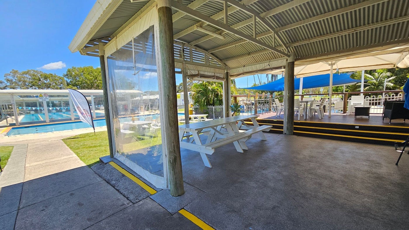 A covered outdoor seating area with a corrugated metal roof and wooden support beams. In the foreground, there are white picnic tables on a concrete floor. To the right, a raised wooden deck with white plastic chairs and tables is shaded by blue and white umbrellas. In the background, a swimming pool with lane markers is visible, surrounded by a grassy area and trees. A clear plastic curtain hangs from the roof, and a flag stands near the pool.