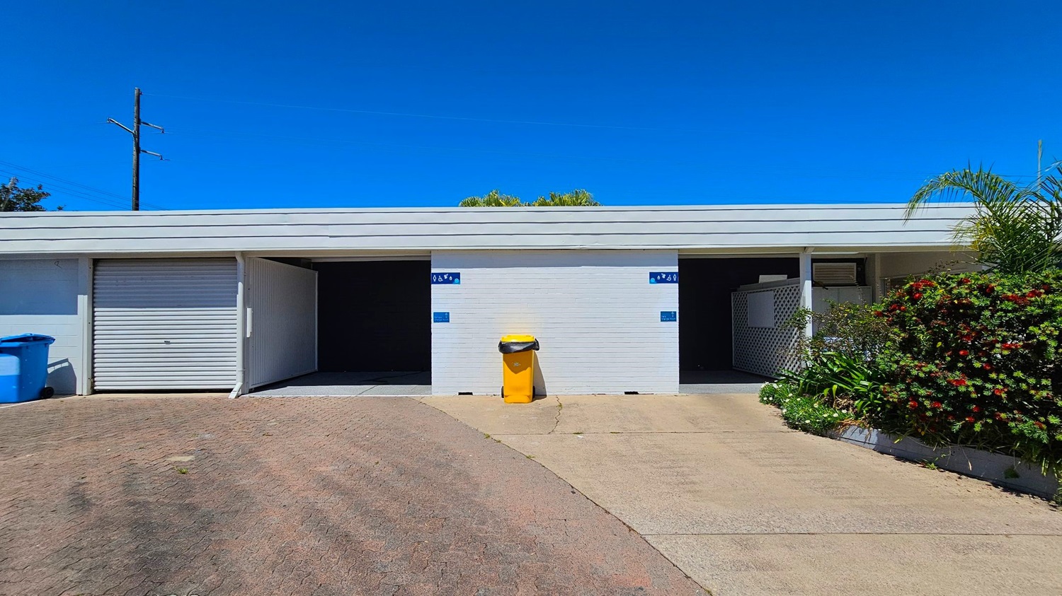 A white building with three sections. On the left, a closed roller door with a blue bin beside it. In the middle, an open garage with a yellow bin in front. On the right, another open garage with lattice panels and plants. The foreground has a paved driveway, and the background shows a clear blue sky and some greenery.