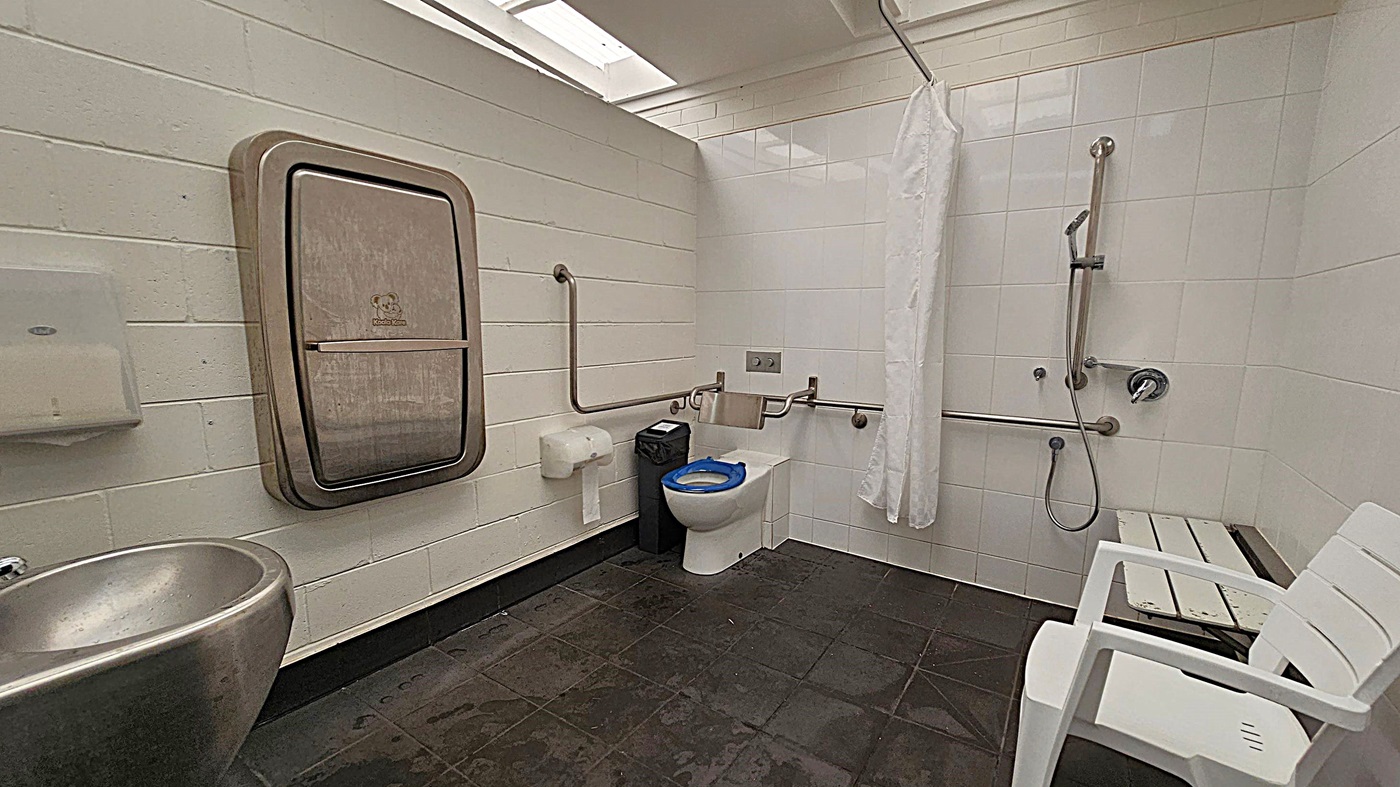 A public restroom with a baby changing station on the left wall, a stainless steel sink below it, and a paper towel dispenser next to it. In the middle, there is a toilet with a blue seat cover, grab bars, a toilet paper holder, and a waste bin. On the right, there is a shower area with a handheld showerhead, grab bars, a white shower curtain, and a white plastic chair. The floor is tiled in dark grey, and the walls are tiled in light colours. The ceiling has a skylight.