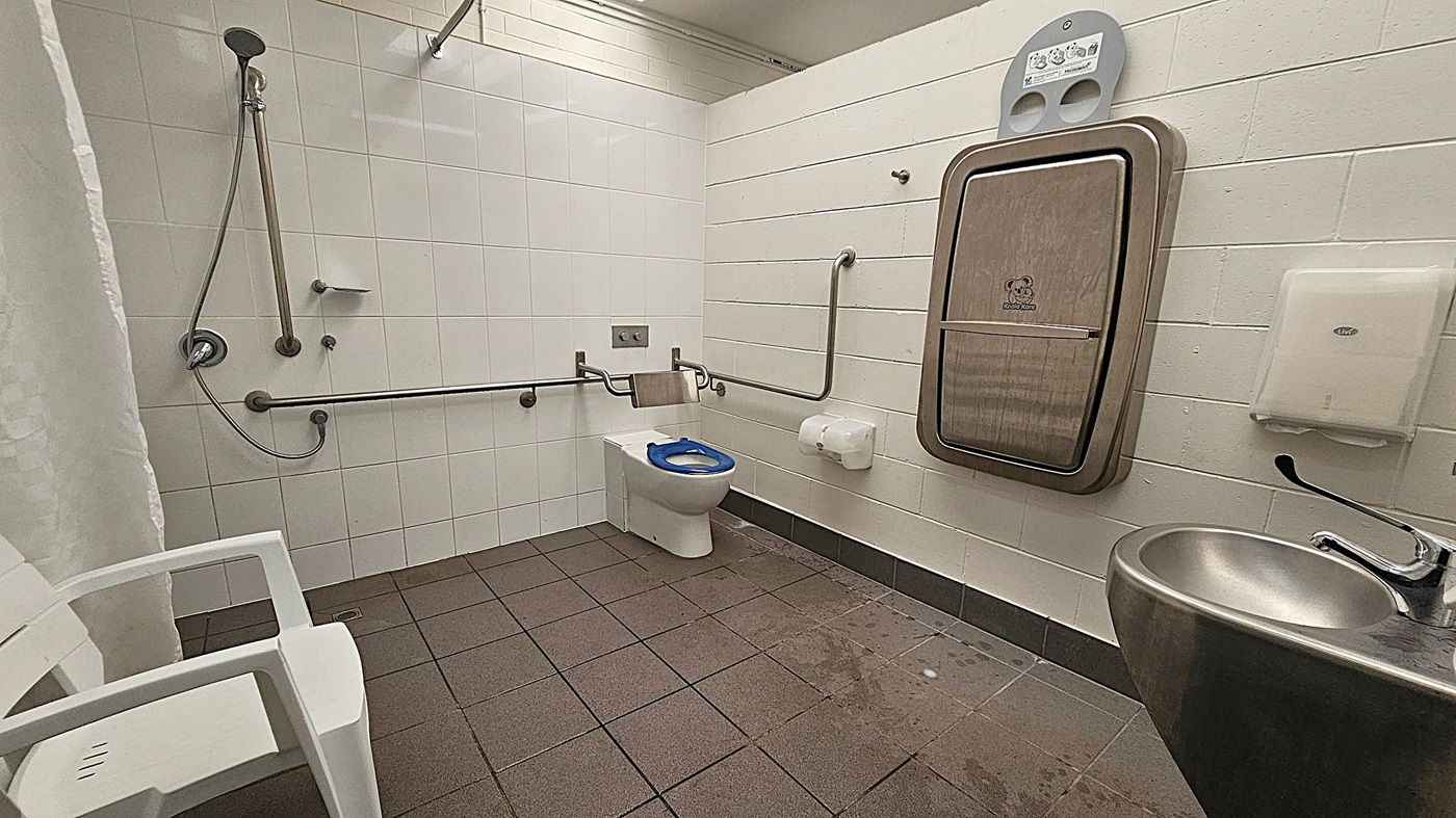 A spacious, accessible bathroom with light green tiled walls and a brown tiled floor. On the left, there is a shower area with a handheld showerhead and a white plastic chair. In the middle, there is a toilet with a blue seat and multiple grab bars around it. On the right, there is a baby changing station mounted on the wall, a paper towel dispenser, and a stainless steel sink with a lever handle.