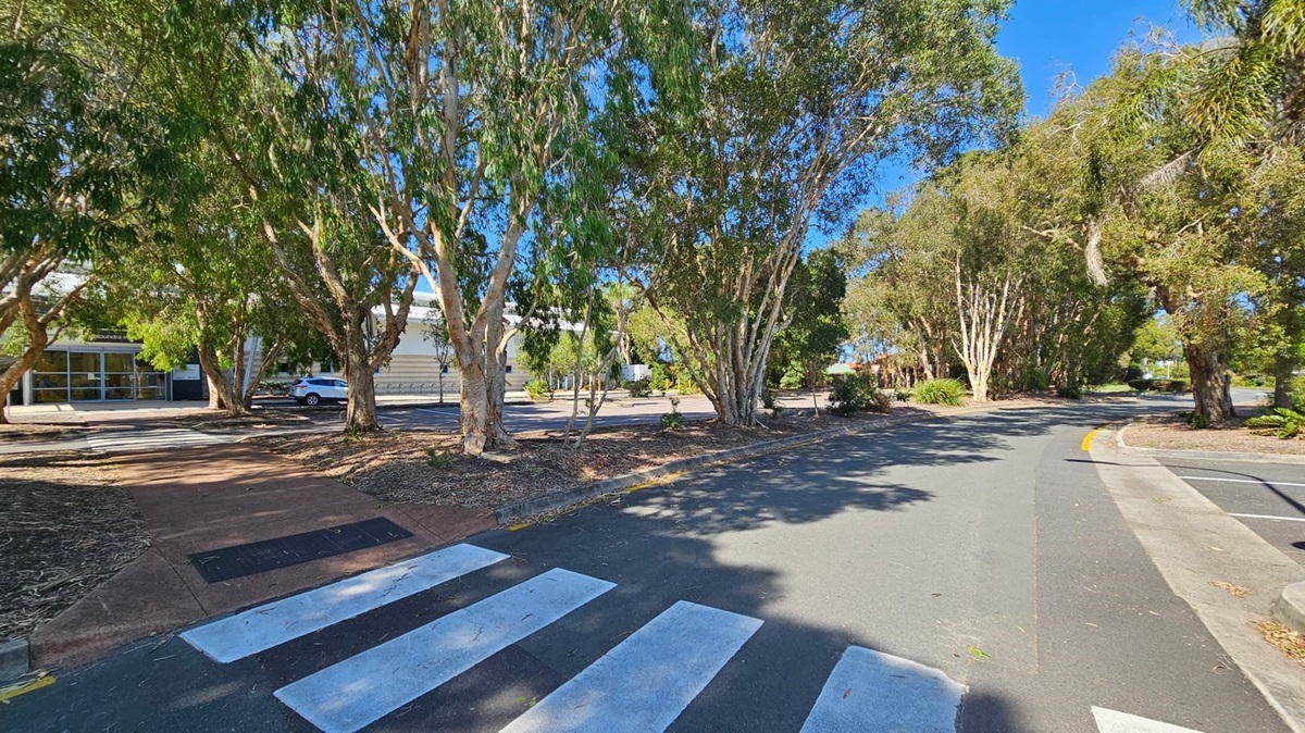 A zebra crossing leads to a path surrounded by tall trees with dense foliage. On the left, a building with large windows and a parked car is visible. The road curves to the right, lined with more trees and some low shrubs. The ground is a mix of asphalt and mulch, with shadows cast by the trees. The sky is clear and blue. A zebra crossing leads to a path surrounded by tall trees with dense foliage. On the left, a building with large windows and a parked car is visible. The road curves to the right, lined with more trees and some low shrubs. The ground is a mix of asphalt and mulch, with shadows cast by the trees. The sky is clear and blue.