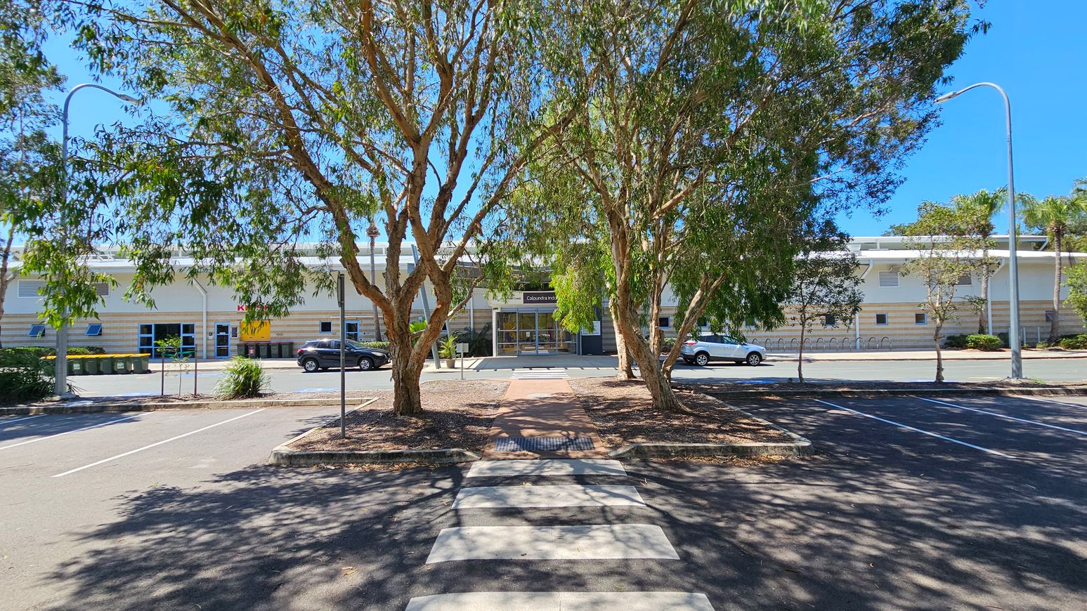A car park with a zebra crossing in the foreground leads to a building in the background. The building has a modern design with a glass entrance and a sign above the door. Two large trees with green foliage are in the centre, casting shadows on the ground. Cars are parked along the street in front of the building. Street lamps are visible on both sides, and there are several bins to the left. The sky is clear and blue. A car park with a zebra crossing in the foreground leads to a building in the background. The building has a modern design with a glass entrance and a sign above the door. Two large trees with green foliage are in the centre, casting shadows on the ground. Cars are parked along the street in front of the building. Street lamps are visible on both sides, and there are several bins to the left. The sky is clear and blue.