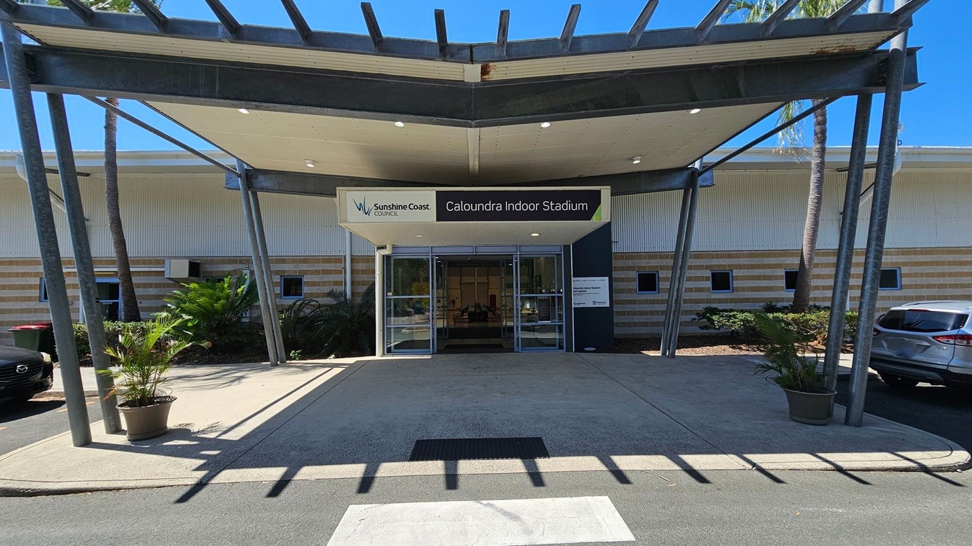 Entrance to Caloundra Indoor Stadium with a sign above the glass doors. The building has a striped exterior with small square windows. A metal awning supported by angled beams extends over the entrance. Potted plants are on either side of the walkway. Cars are parked on the right, and there are palm trees in the background. The ground is paved, and there is a pedestrian crossing in the foreground. Entrance to Caloundra Indoor Stadium with a sign above the glass doors. The building has a striped exterior with small square windows. A metal awning supported by angled beams extends over the entrance. Potted plants are on either side of the walkway. Cars are parked on the right, and there are palm trees in the background. The ground is paved, and there is a pedestrian crossing in the foreground.