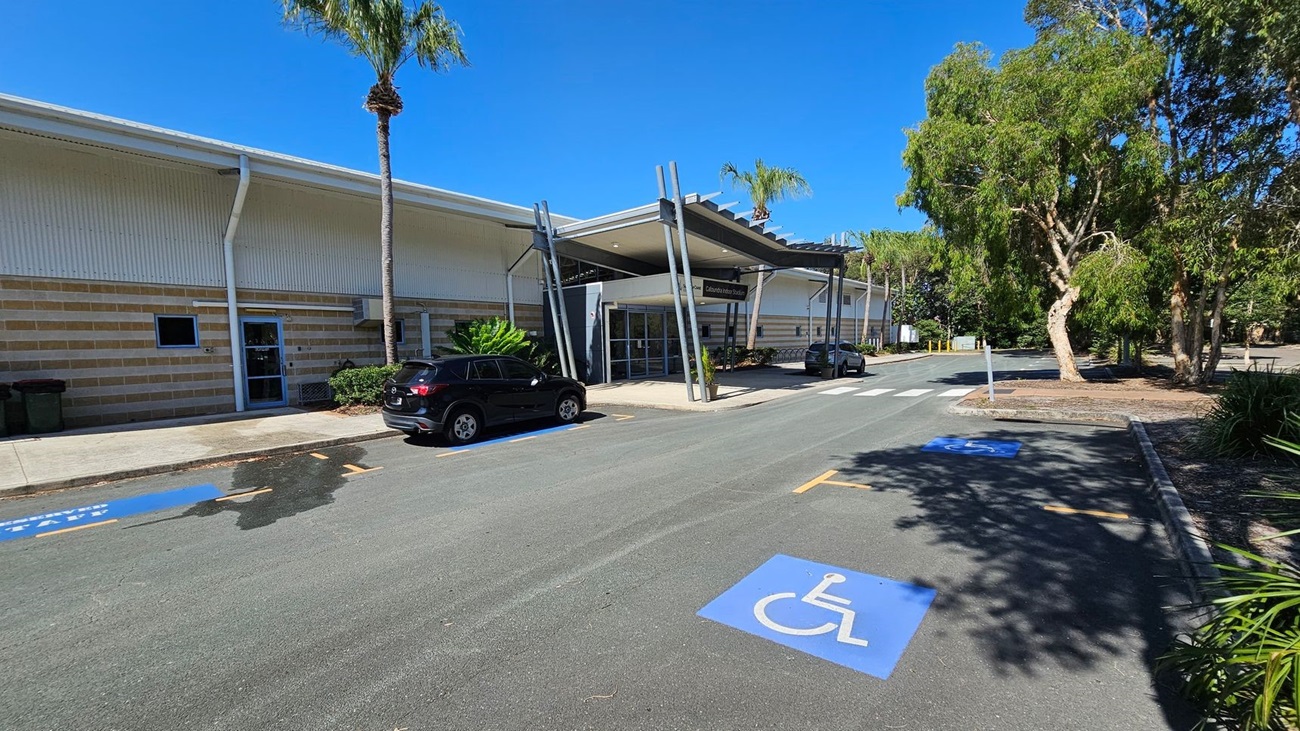 A modern building with a striped exterior and a large entrance canopy supported by angled metal beams. On the left, a black car is parked near a small palm tree. The foreground features a marked disabled parking space on the asphalt. On the right, there are several large trees and landscaped greenery. The sky is clear and blue. A modern building with a striped exterior and a large entrance canopy supported by angled metal beams. On the left, a black car is parked near a small palm tree. The foreground features a marked disabled parking space on the asphalt. On the right, there are several large trees and landscaped greenery. The sky is clear and blue.