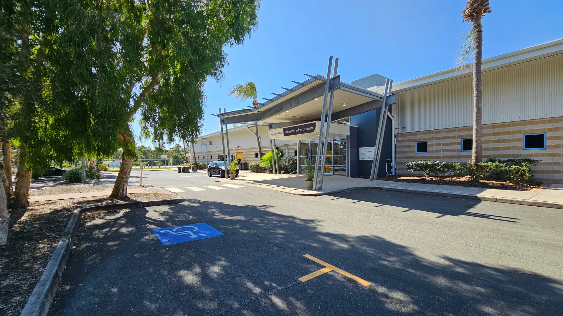 A large building with a modern entrance labelled "Caloundra Indoor Stadium" is on the right. The structure has a metal canopy supported by angled beams. A car is parked near the entrance. On the left, there are several large trees casting shadows on the ground. The pavement has a blue disabled parking symbol. The building's exterior is a mix of beige and cream with small windows. A palm tree is visible near the entrance. A large building with a modern entrance labelled "Caloundra Indoor Stadium" is on the right. The structure has a metal canopy supported by angled beams. A car is parked near the entrance. On the left, there are several large trees casting shadows on the ground. The pavement has a blue disabled parking symbol. The building's exterior is a mix of beige and cream with small windows. A palm tree is visible near the entrance.