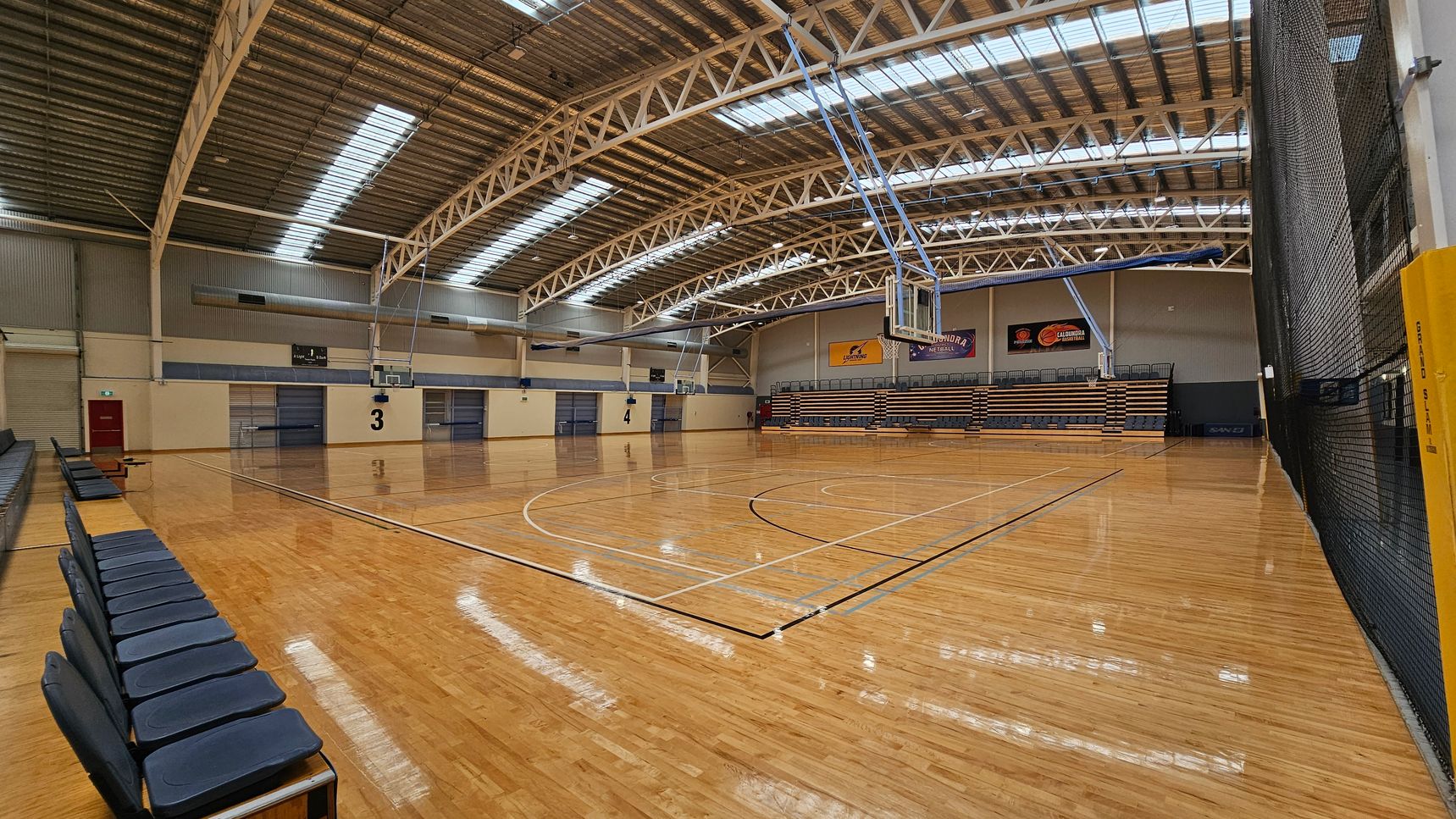 A large indoor sports hall with a polished wooden floor. In the centre, there are two rows of blue chairs facing opposite directions, with "WELCOME" signs at the front. The ceiling features exposed metal beams and skylights. The background shows four basketball courts, each marked with numbers 1 to 4, and basketball hoops. The walls are light-coloured with some banners and doors visible. A large indoor sports hall with a polished wooden floor. In the centre, there are two rows of blue chairs facing opposite directions, with "WELCOME" signs at the front. The ceiling features exposed metal beams and skylights. The background shows four basketball courts, each marked with numbers 1 to 4, and basketball hoops. The walls are light-coloured with some banners and doors visible.