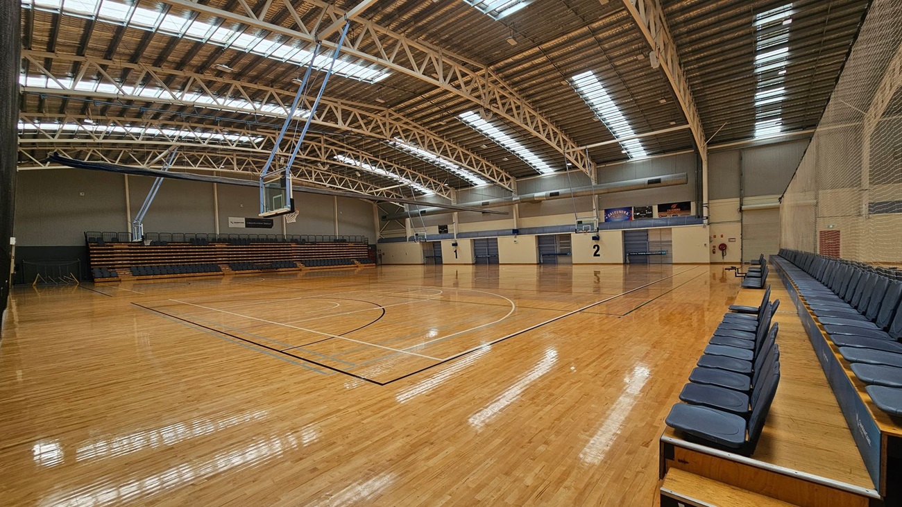 A spacious indoor basketball court with a polished wooden floor. On the left, there are retractable bleachers with dark seating. The right side features more seating along the wall. The ceiling is high with exposed metal beams and skylights, allowing natural light to illuminate the space. Two basketball hoops are visible, one on each end of the court. The walls are light-coloured with some signage and markings indicating court numbers. A spacious indoor basketball court with a polished wooden floor. On the left, there are retractable bleachers with dark seating. The right side features more seating along the wall. The ceiling is high with exposed metal beams and skylights, allowing natural light to illuminate the space. Two basketball hoops are visible, one on each end of the court. The walls are light-coloured with some signage and markings indicating court numbers.