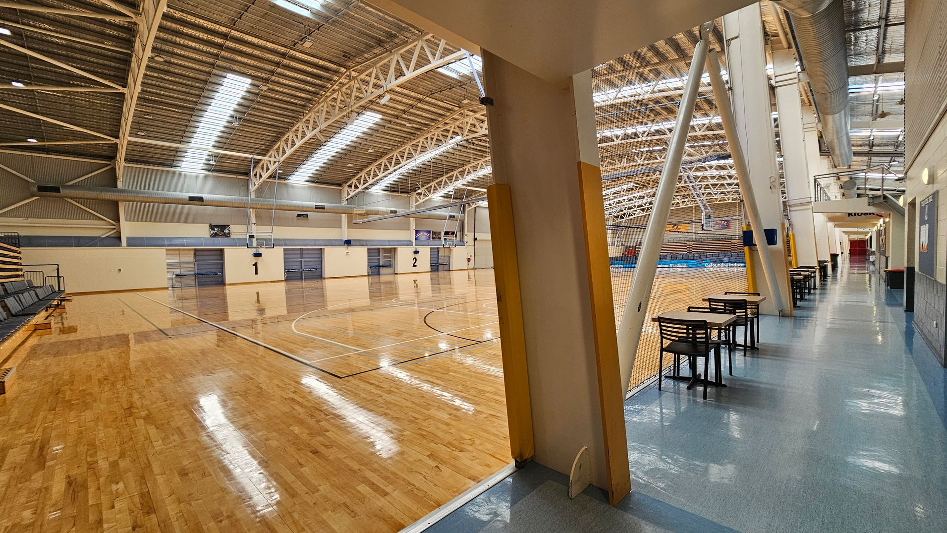 A large indoor sports hall with a polished wooden floor on the left, featuring two basketball courts marked with numbers 1 and 2. The ceiling is high with metal beams and skylights. On the right, a corridor with a blue floor has tables and chairs lined up against a netted barrier. The corridor leads to a distant area marked "Kiosk." The walls are light-coloured, and there are exposed ducts and structural supports visible. A large indoor sports hall with a polished wooden floor on the left, featuring two basketball courts marked with numbers 1 and 2. The ceiling is high with metal beams and skylights. On the right, a corridor with a blue floor has tables and chairs lined up against a netted barrier. The corridor leads to a distant area marked "Kiosk." The walls are light-coloured, and there are exposed ducts and structural supports visible.