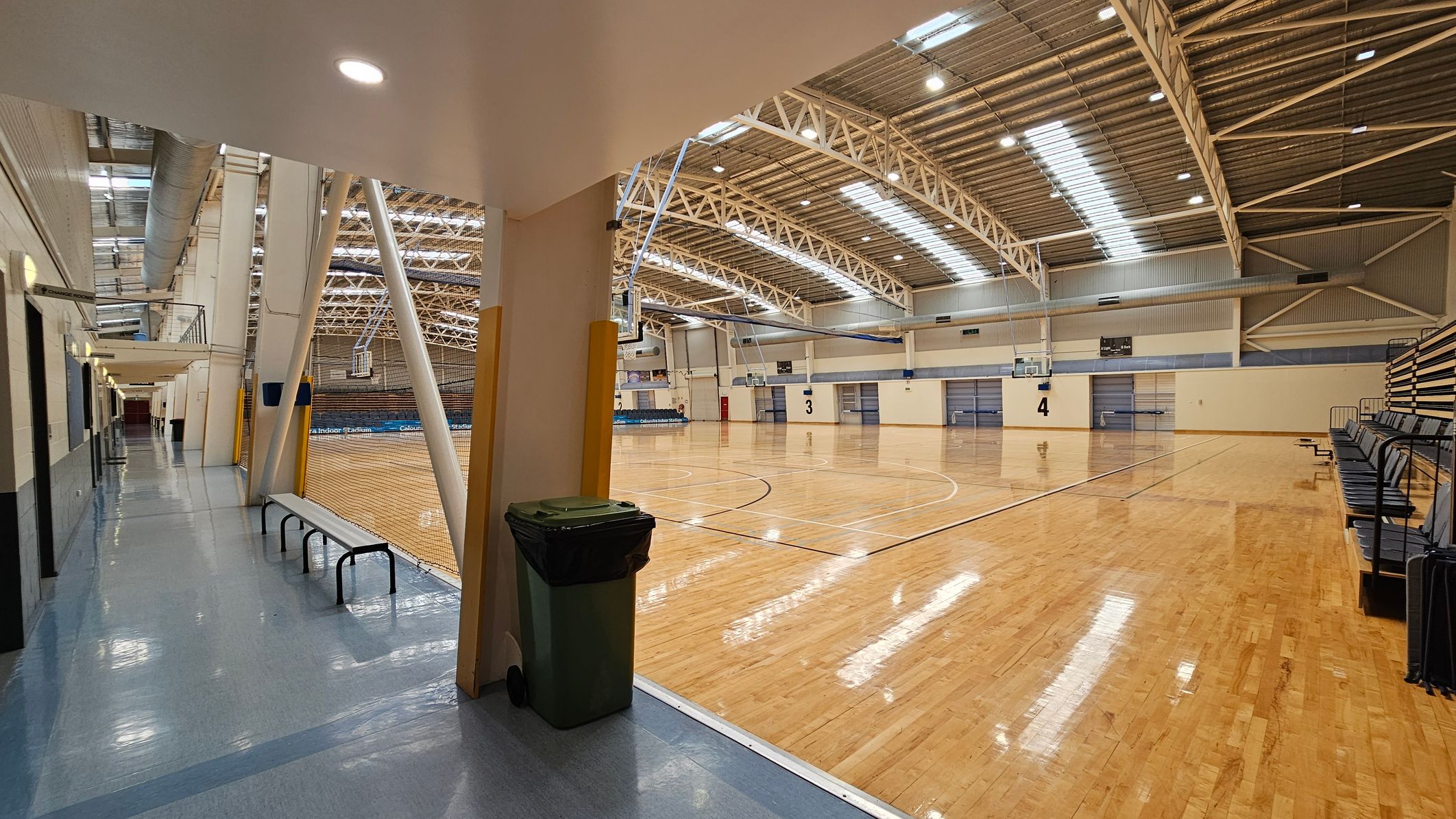 A large indoor sports hall with a polished wooden floor and high, metal truss ceiling. On the right, there are rows of retractable bleachers. In the centre, basketball hoops are visible, and the court is marked with lines. On the left, a corridor with a shiny blue floor leads to the hall, featuring a bench and a rubbish bin. The ceiling has skylights and industrial lighting. A large indoor sports hall with a polished wooden floor and high, metal truss ceiling. On the right, there are rows of retractable bleachers. In the centre, basketball hoops are visible, and the court is marked with lines. On the left, a corridor with a shiny blue floor leads to the hall, featuring a bench and a rubbish bin. The ceiling has skylights and industrial lighting.