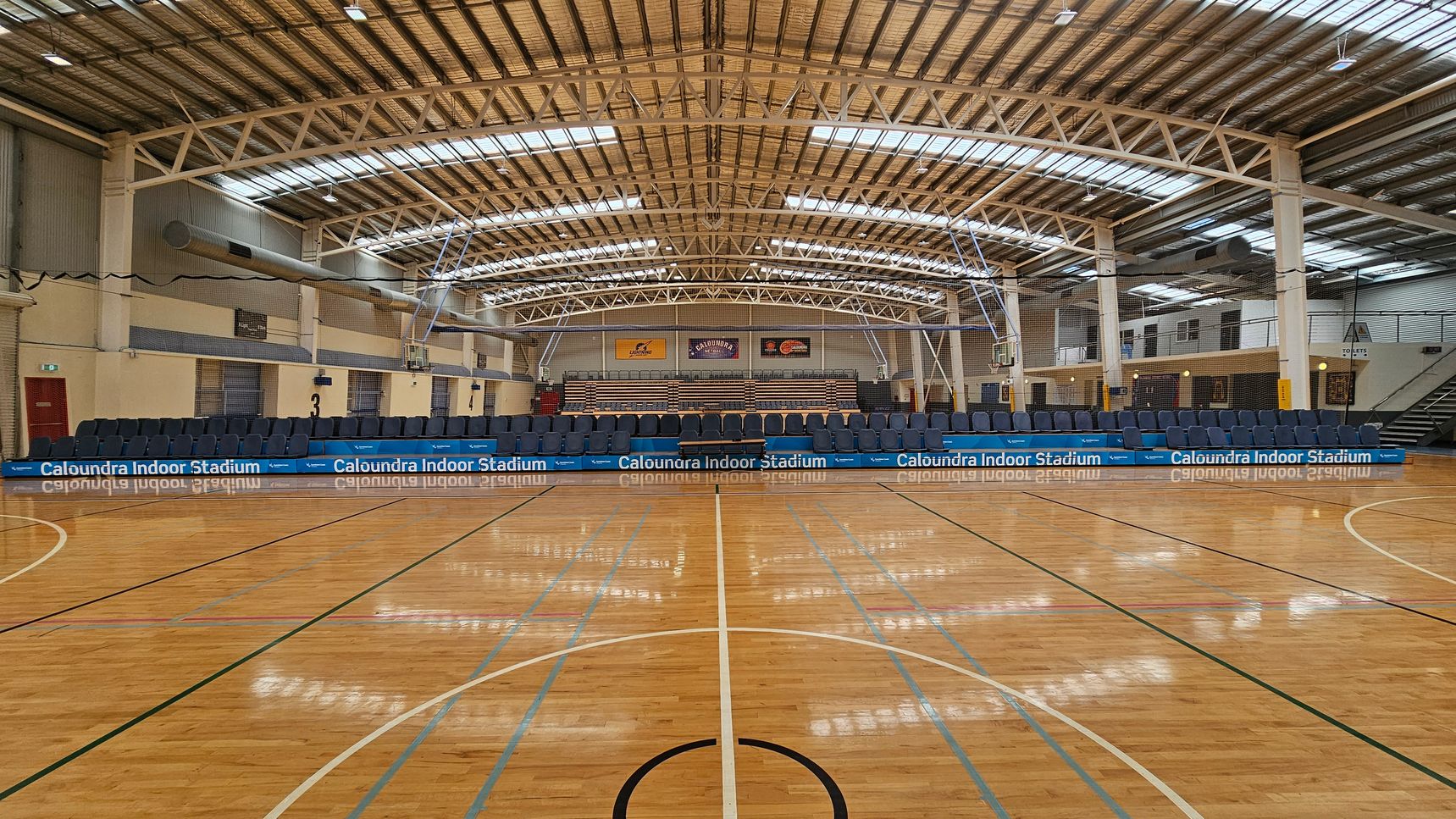 A spacious indoor sports stadium with a polished wooden floor marked for basketball. In the background, there are rows of blue seats and a scoreboard. The ceiling is high with metal beams and skylights, allowing natural light to illuminate the space. Banners are displayed on the far wall above the seating area. The walls are a mix of brick and metal panelling, and there are doors and staircases on the right side. A spacious indoor sports stadium with a polished wooden floor marked for basketball. In the background, there are rows of blue seats and a scoreboard. The ceiling is high with metal beams and skylights, allowing natural light to illuminate the space. Banners are displayed on the far wall above the seating area. The walls are a mix of brick and metal panelling, and there are doors and staircases on the right side.