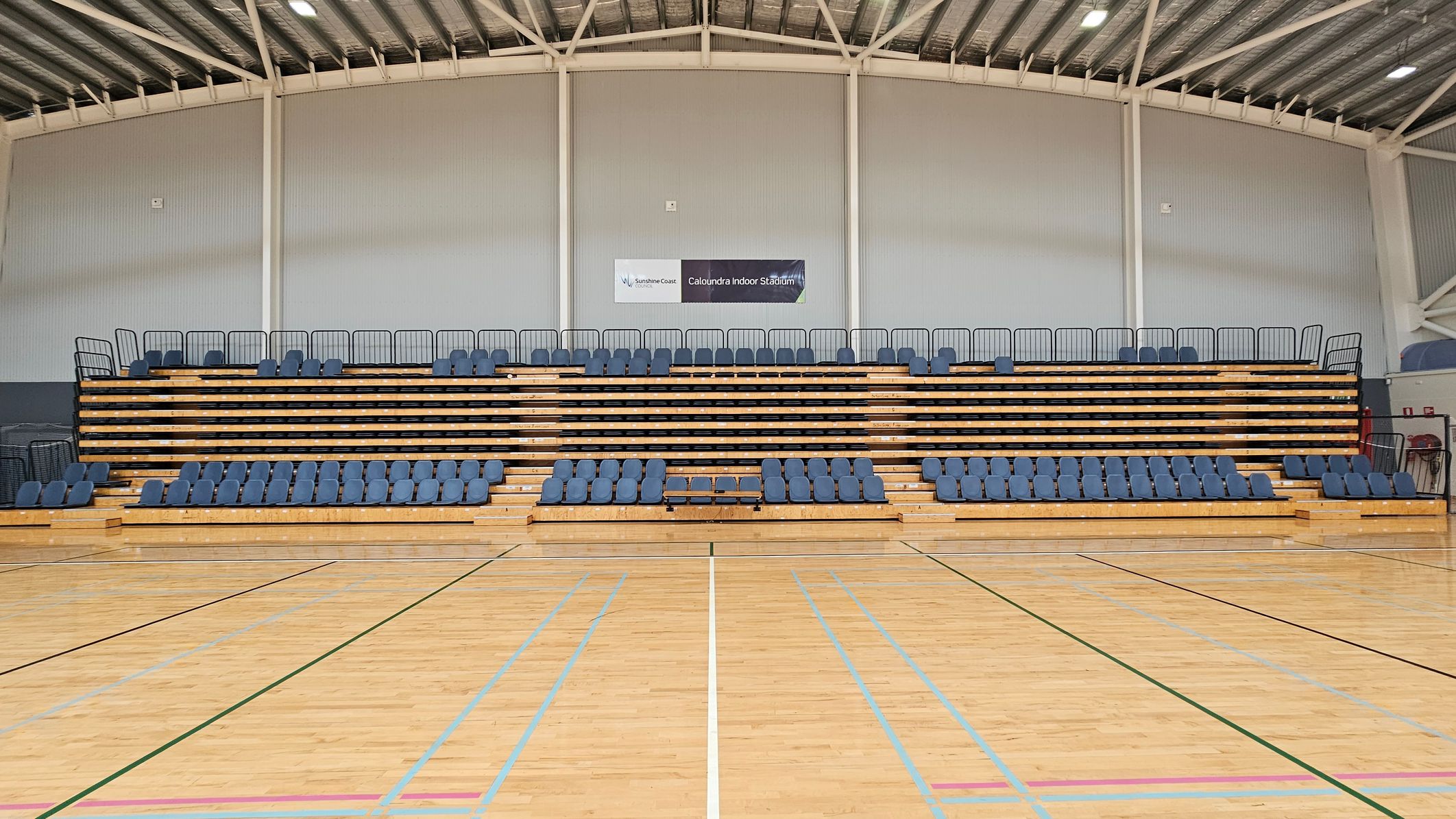 Indoor sports stadium with wooden bleachers featuring blue seats. The bleachers are centred against a grey wall with a sign reading "Caloundra Indoor Stadium" above. The floor is a polished wooden sports court with various coloured lines. The ceiling is high with exposed beams and lighting fixtures. Indoor sports stadium with wooden bleachers featuring blue seats. The bleachers are centred against a grey wall with a sign reading "Caloundra Indoor Stadium" above. The floor is a polished wooden sports court with various coloured lines. The ceiling is high with exposed beams and lighting fixtures.