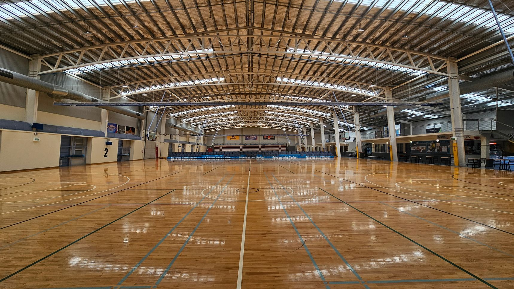 A spacious indoor sports stadium with a polished wooden floor marked for basketball. In the background, there are rows of blue seats and a scoreboard. The ceiling is high with metal beams and skylights, allowing natural light to illuminate the space. Banners are displayed on the far wall above the seating area. The walls are a mix of brick and metal panelling, and there are doors and staircases on the right side. A spacious indoor sports stadium with a polished wooden floor marked for basketball. In the background, there are rows of blue seats and a scoreboard. The ceiling is high with metal beams and skylights, allowing natural light to illuminate the space. Banners are displayed on the far wall above the seating area. The walls are a mix of brick and metal panelling, and there are doors and staircases on the right side.