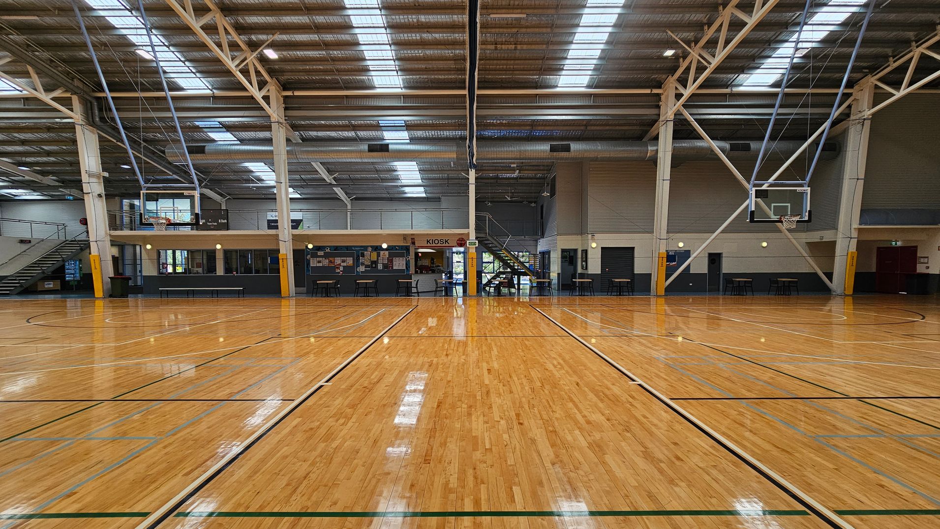 A large indoor sports stadium with a polished wooden floor marked for basketball. The ceiling is high with exposed beams and skylights. On the left, there are numbered walls and a basketball hoop. On the right, there is a kiosk area with tables and chairs. In the background, rows of blue seating are visible, along with banners and signs. The space is well-lit with natural light from the skylights. A large indoor sports stadium with a polished wooden floor marked for basketball. The ceiling is high with exposed beams and skylights. On the left, there are numbered walls and a basketball hoop. On the right, there is a kiosk area with tables and chairs. In the background, rows of blue seating are visible, along with banners and signs. The space is well-lit with natural light from the skylights.